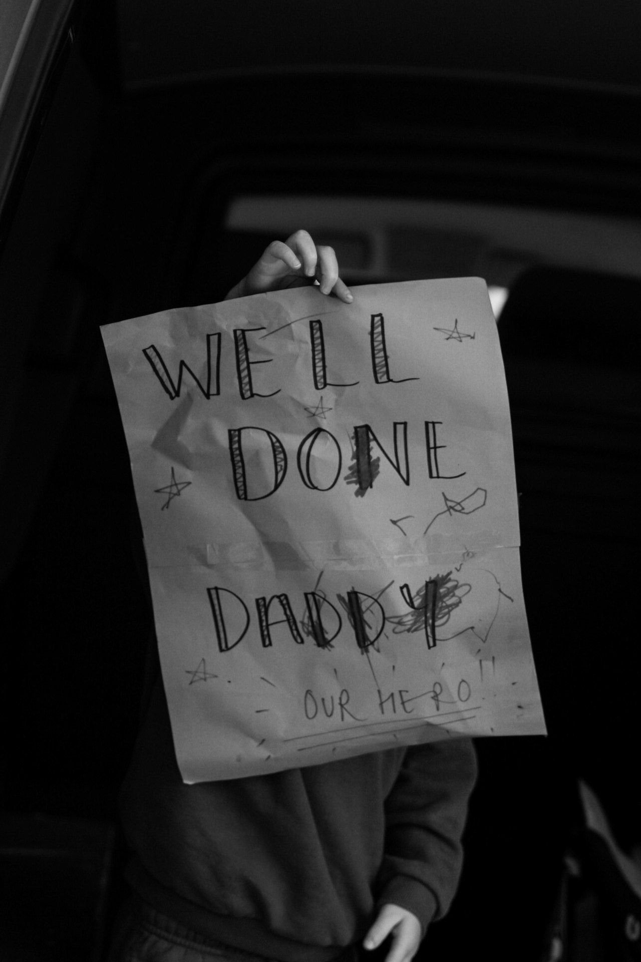 Black and white photo of a child holding a handmade sign reading Well Done Daddy Our Hero.