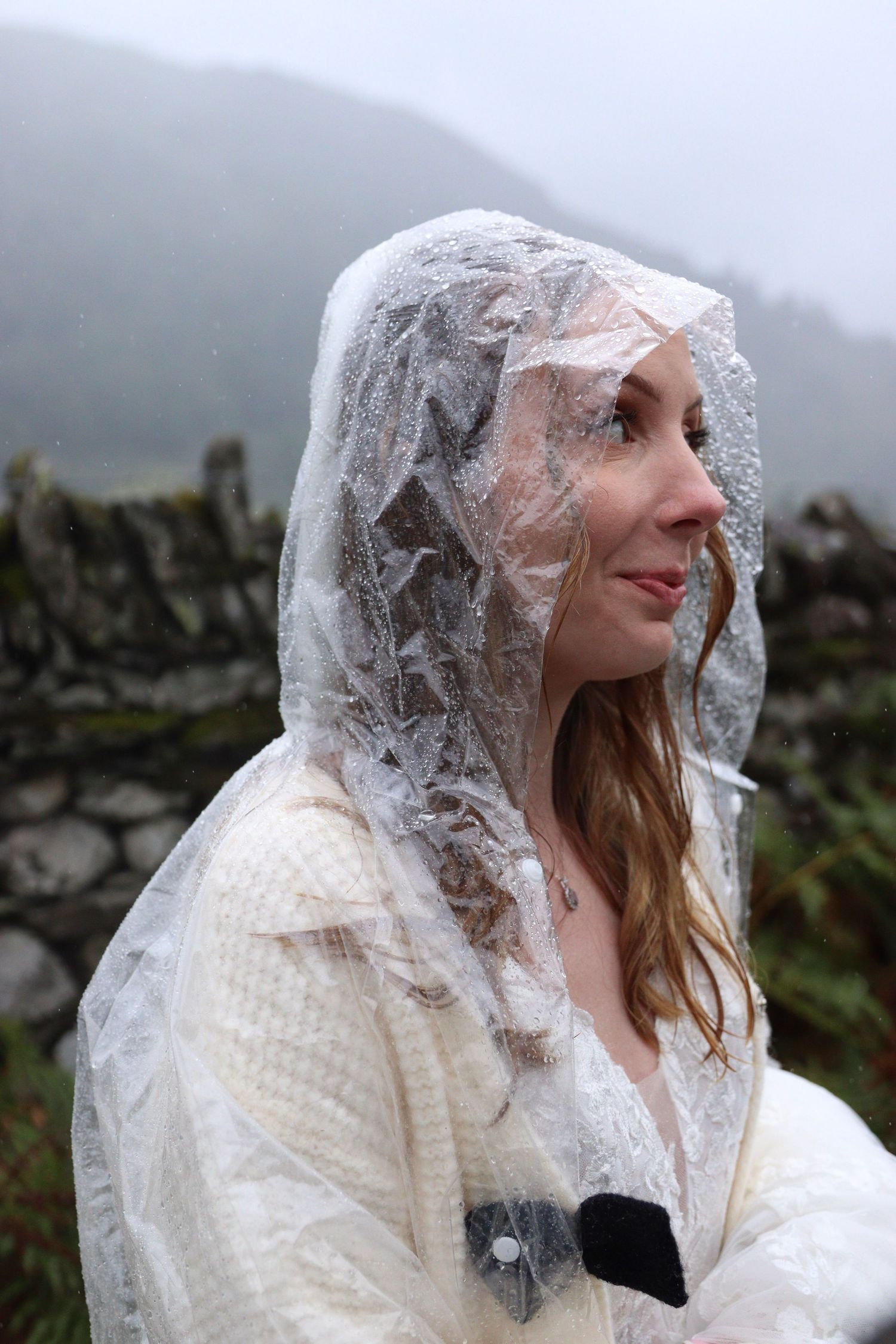 Smiling woman with long auburn hair wears a clear plastic rain poncho over a white dress beside a misty hillside and stone wall.