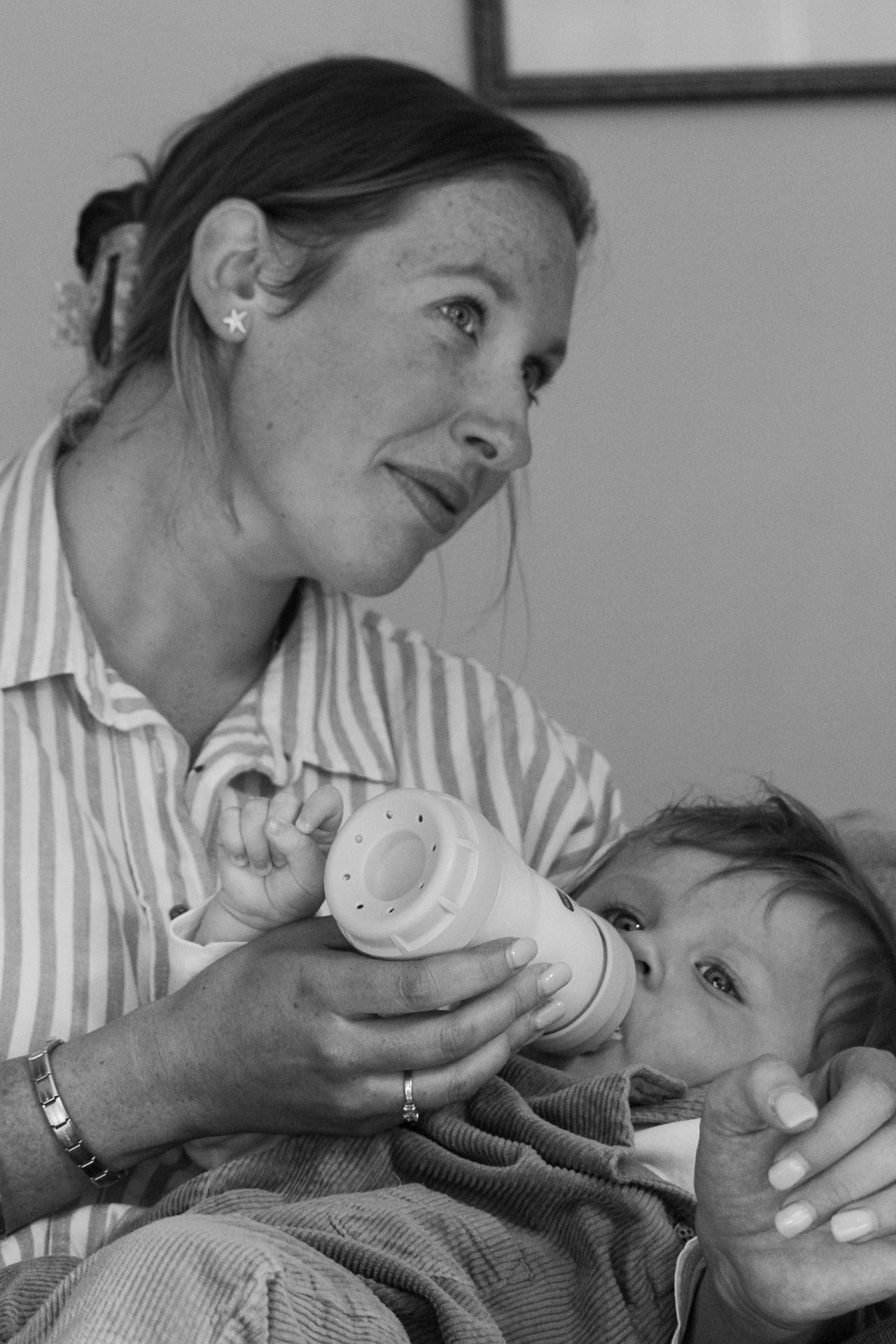 A mother tenderly bottle feeds her infant baby while sitting down, captured in black and white photography.