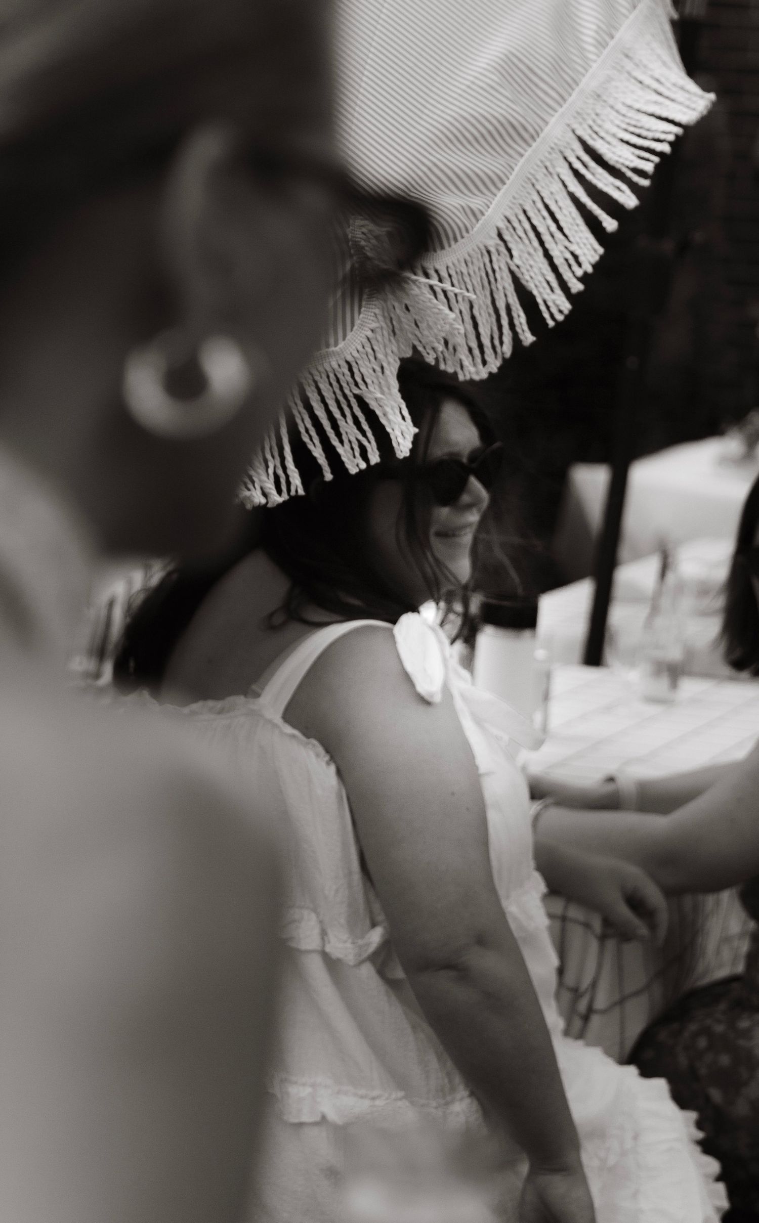 Black and white photo of a smiling woman in sunglasses and fringed hat wearing a white sundress.