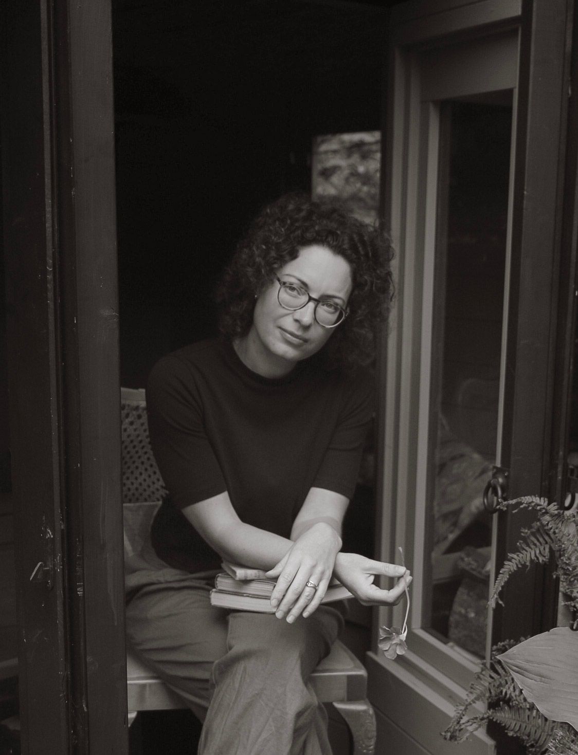 A black and white portrait of a curly-haired woman with glasses sitting in a doorway holding a book.