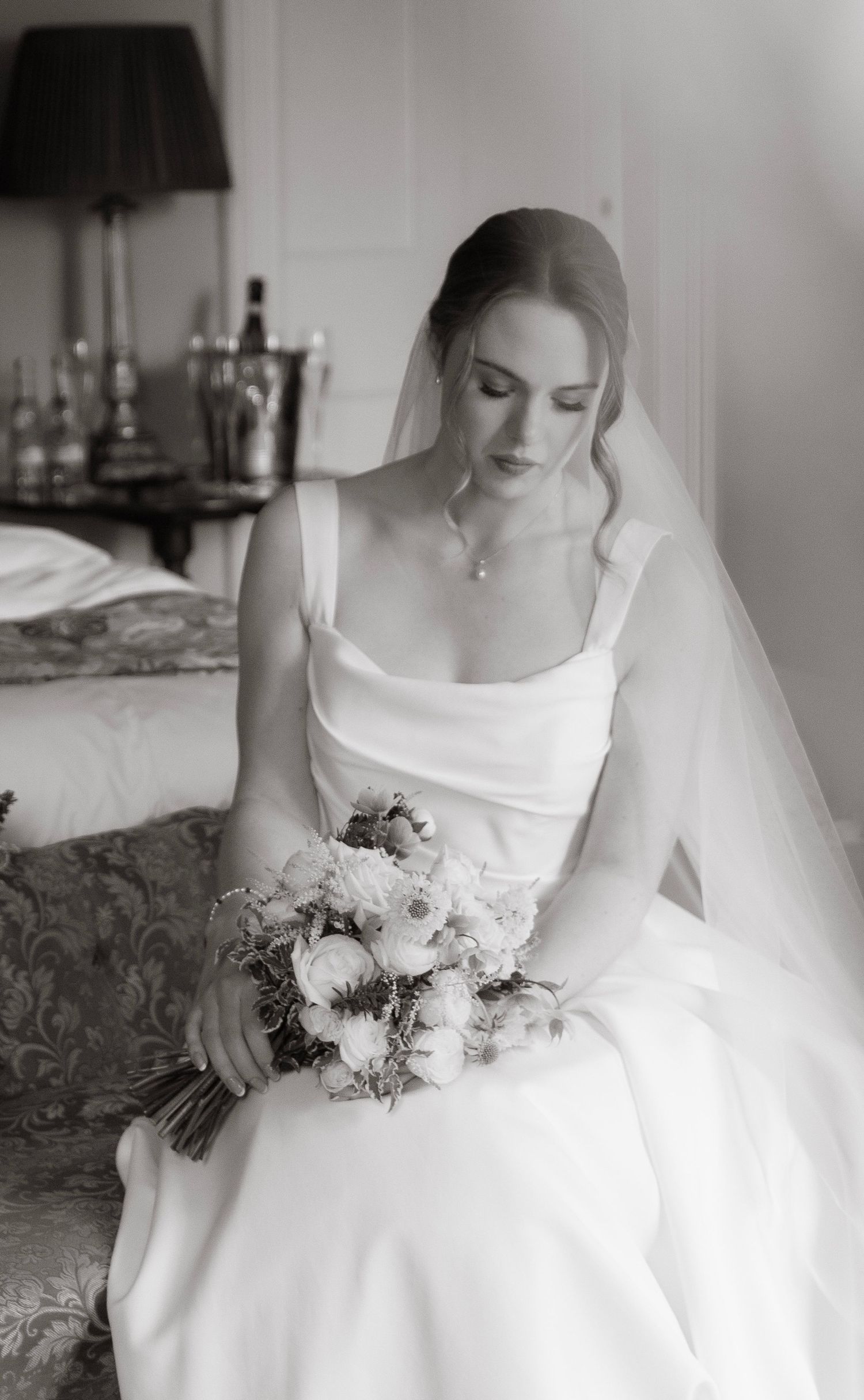 Bride in white gown and veil sits holding a floral bouquet, in a black and white wedding portrait.