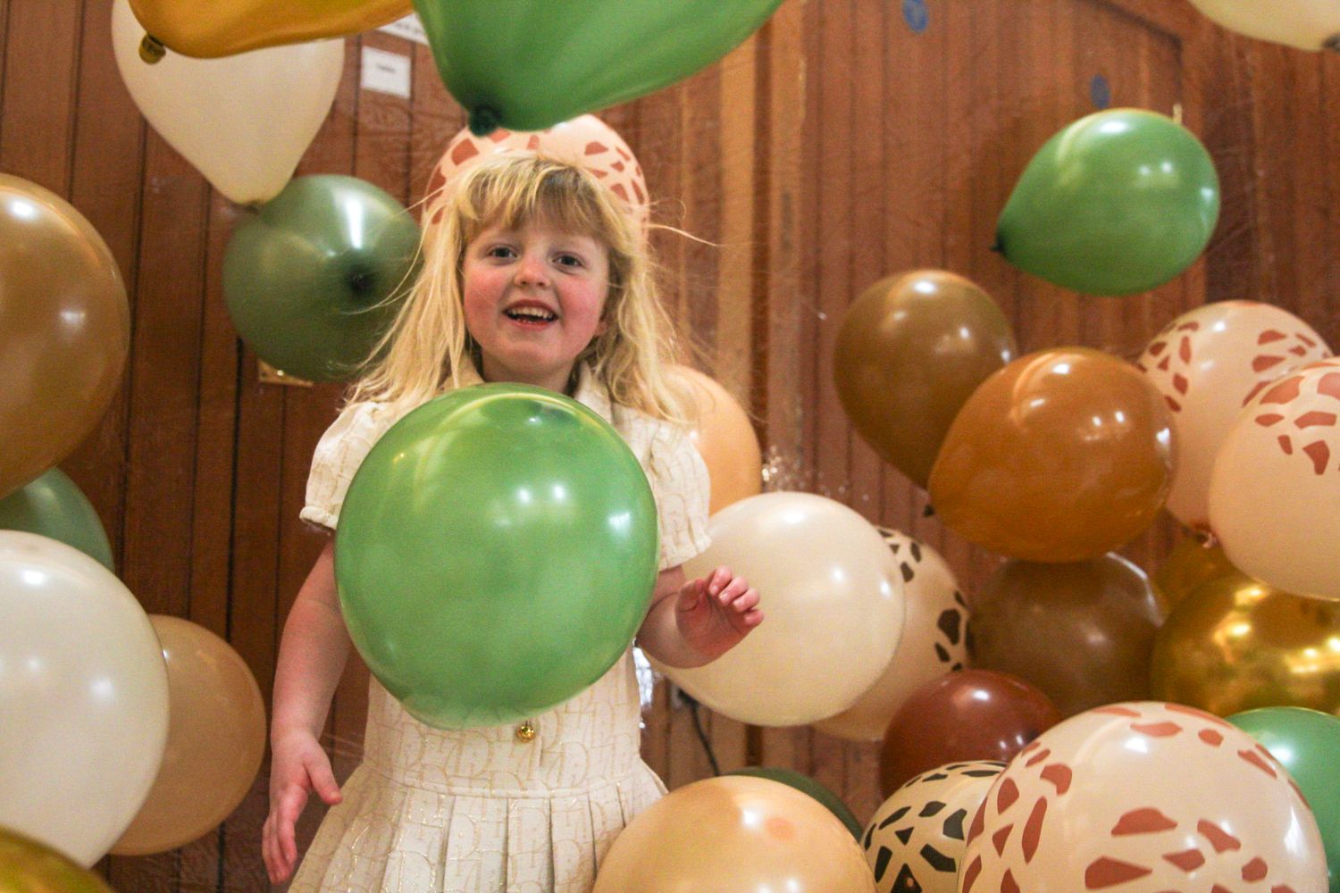 Happy young girl laughing and holding a green balloon surrounded by safari-themed brown and green party balloons.
