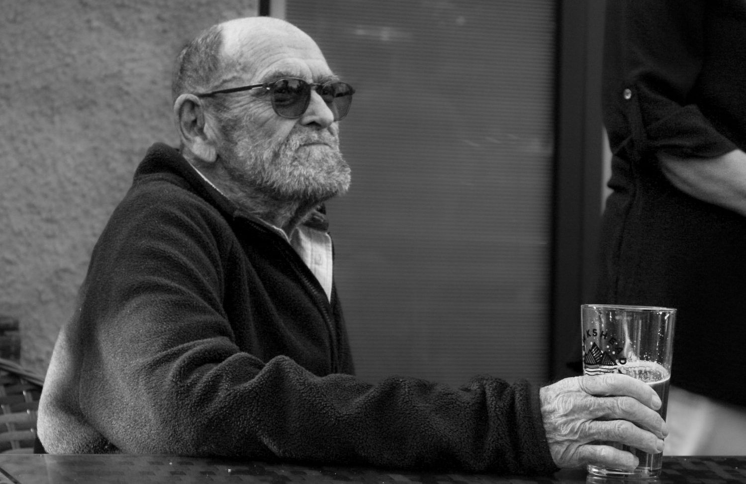 Elderly man with beard and sunglasses holding a drink outdoors in black and white.