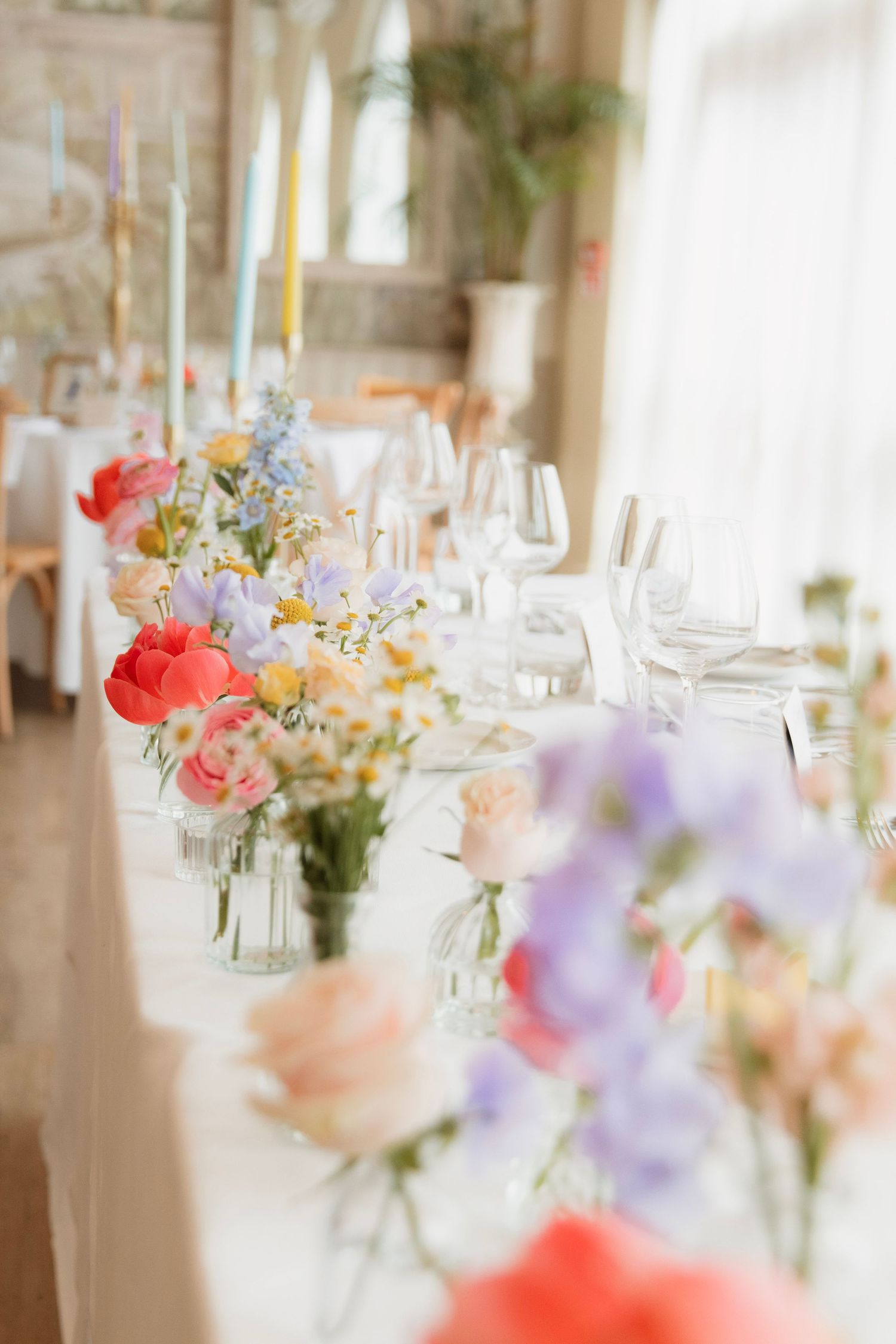 Elegant wedding table with colorful wildflowers in glass vases, pastel candles, and crystal glasses in a bright venue.