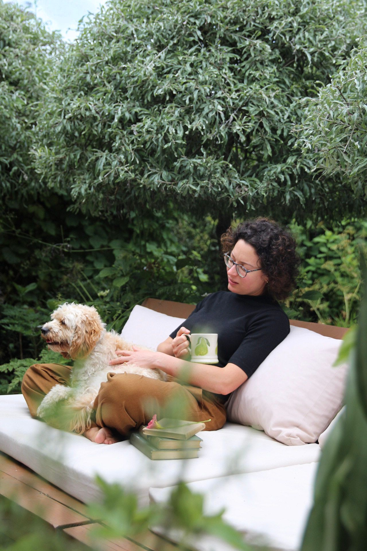 Woman with curly hair relaxing on outdoor sofa with fluffy dog while holding a green mug.