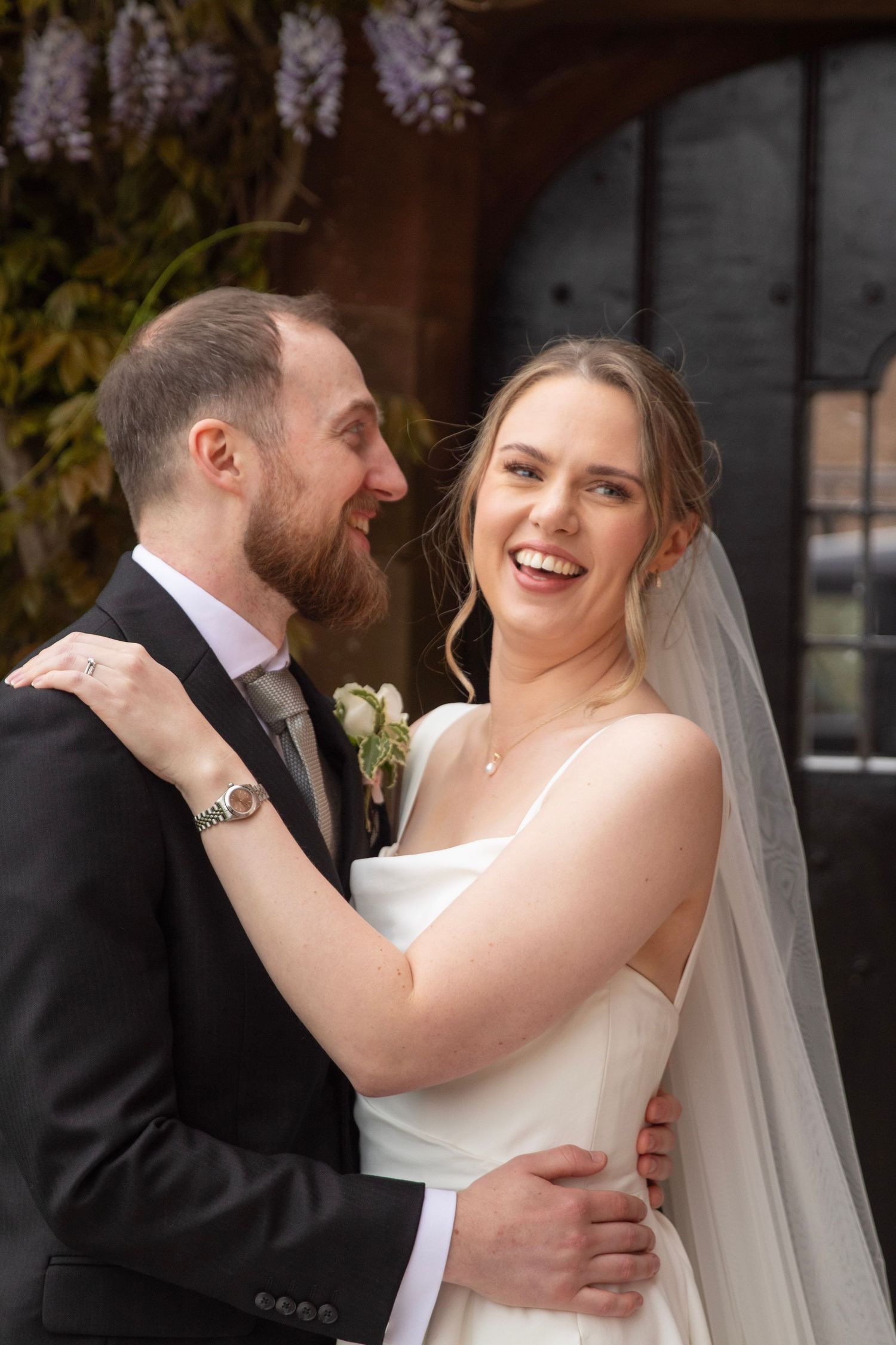 Smiling bride in white gown and veil embraces bearded groom in black suit with boutonniere at wedding venue.