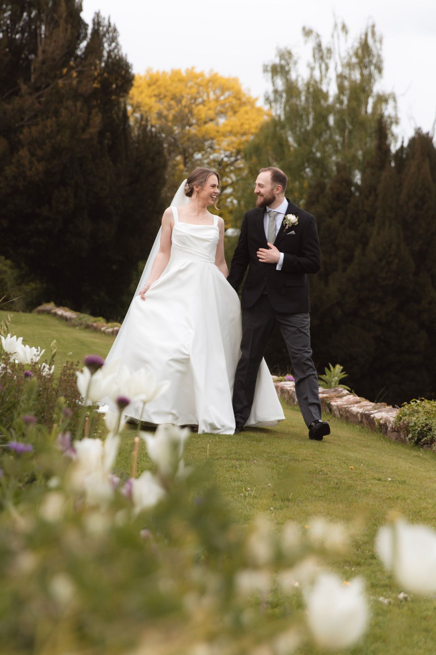 Bride and groom walking hand in hand through a lush garden, smiling at each other on their wedding day.