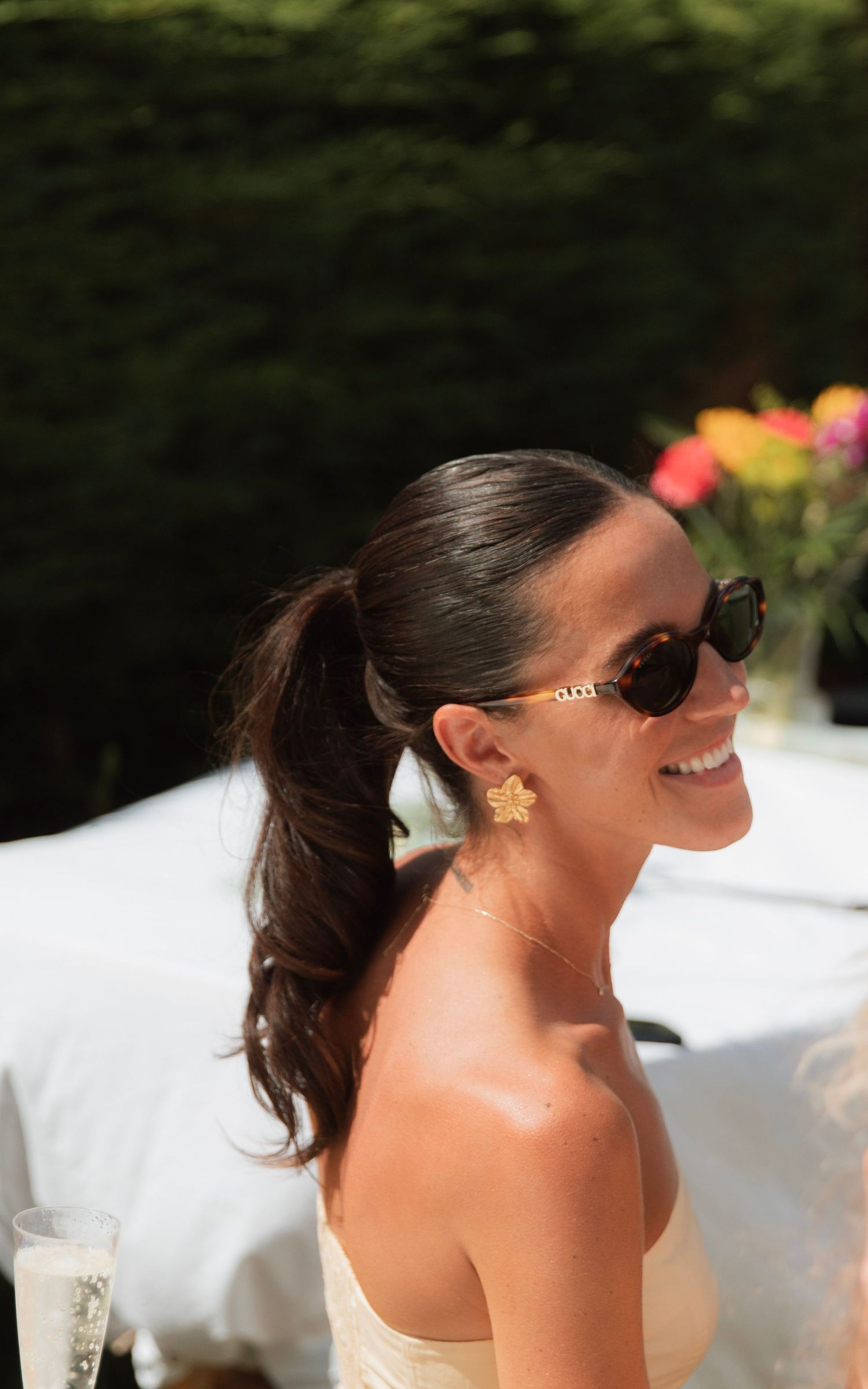 A smiling brunette woman with a ponytail wearing gold flower earrings and sunglasses at an outdoor event.