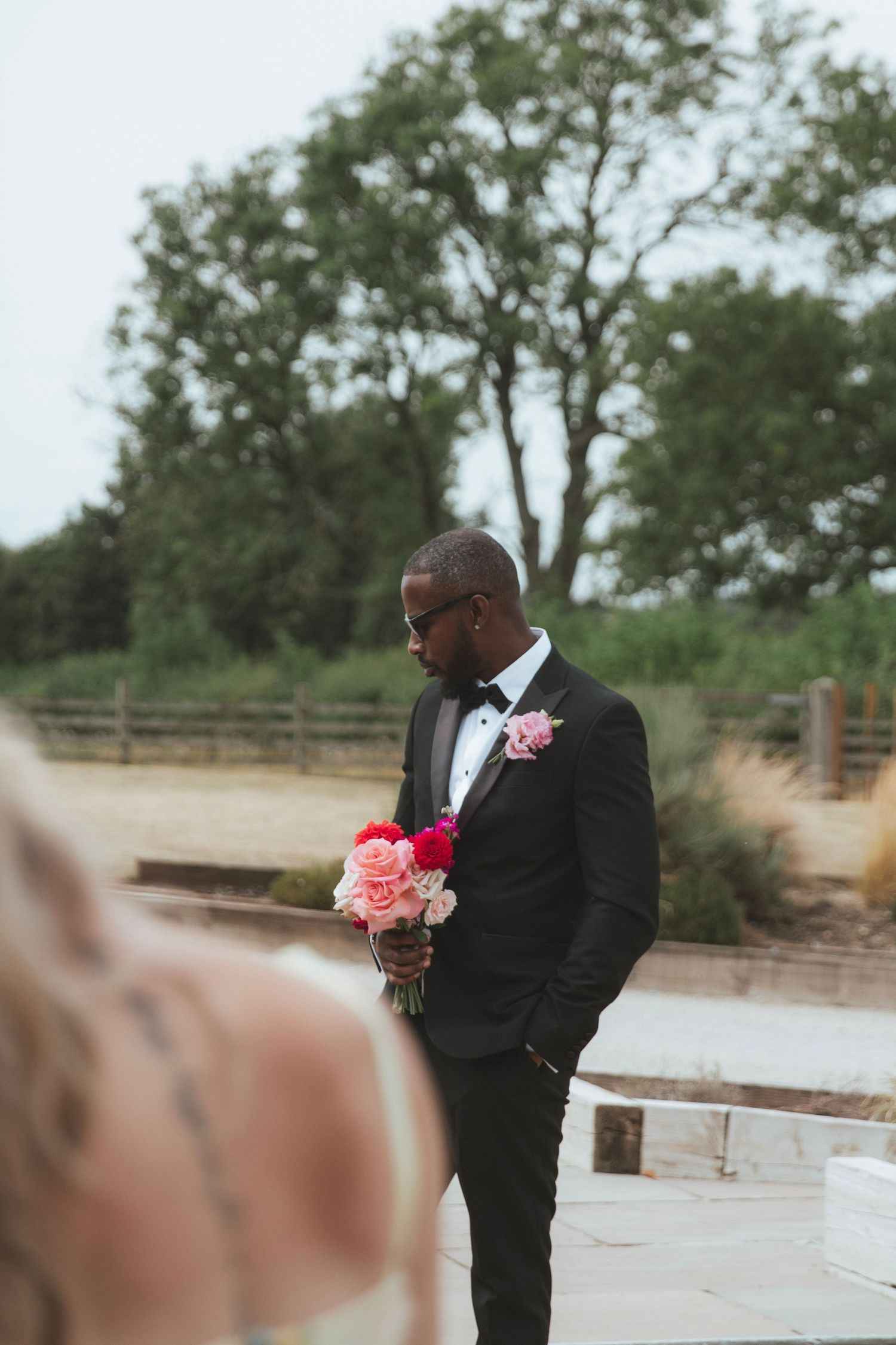 Groom in a black tuxedo and bow tie holds a pink and red bouquet outdoors, a boutonniere on his lapel, with trees in the background.