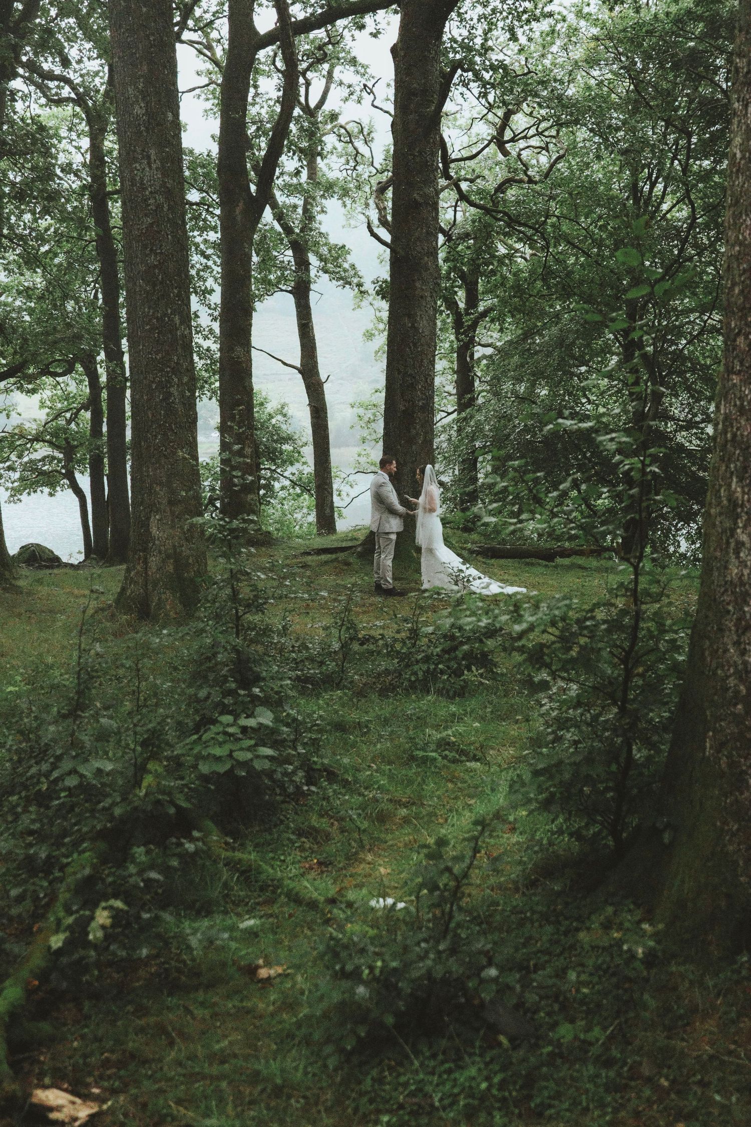 Bride in a long white veil and groom standing together in a misty, moss-covered forest clearing during an intimate woodland elopement.