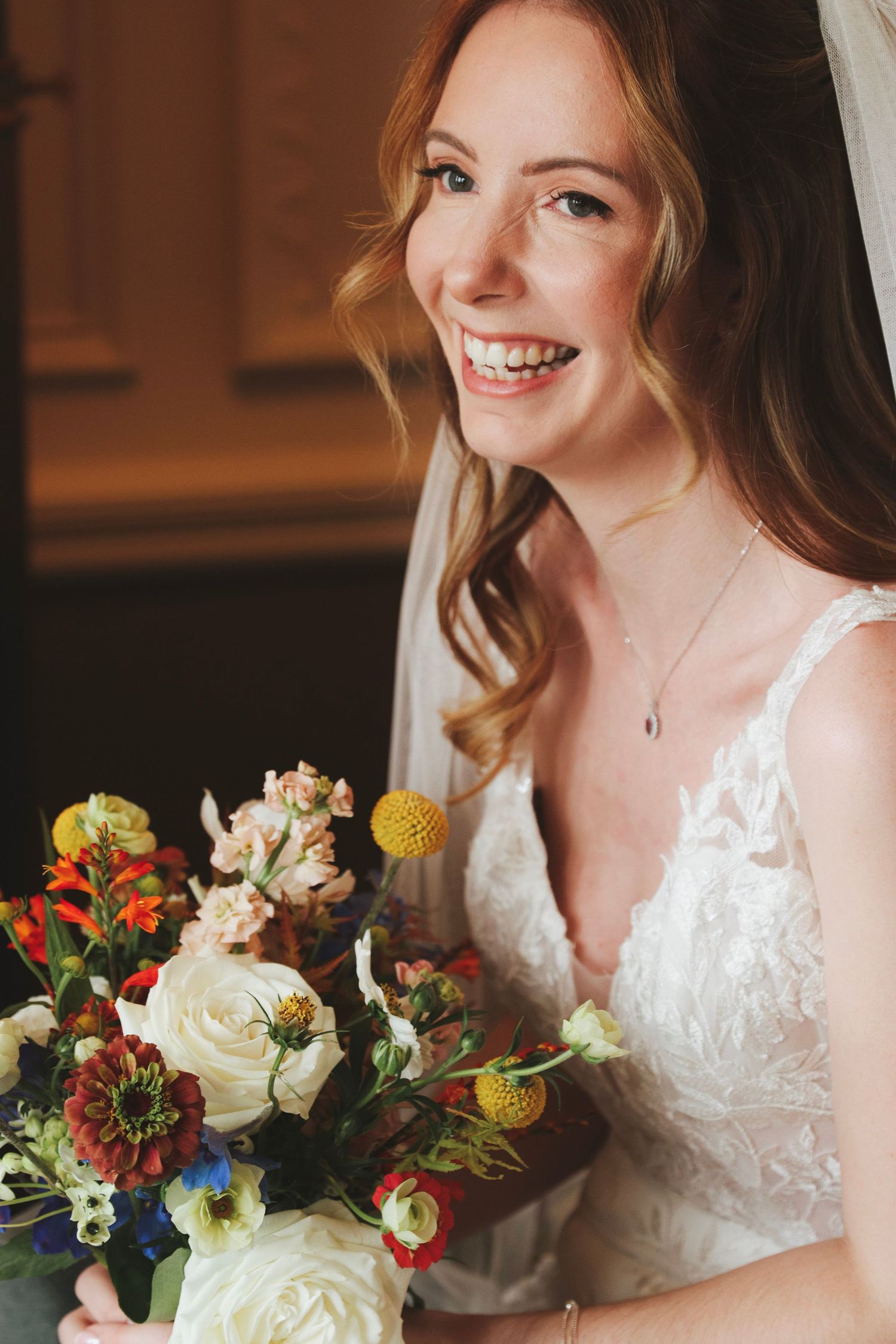 Smiling bride in a white lace wedding dress and veil holding a colorful bouquet of roses, marigolds and wildflowers.