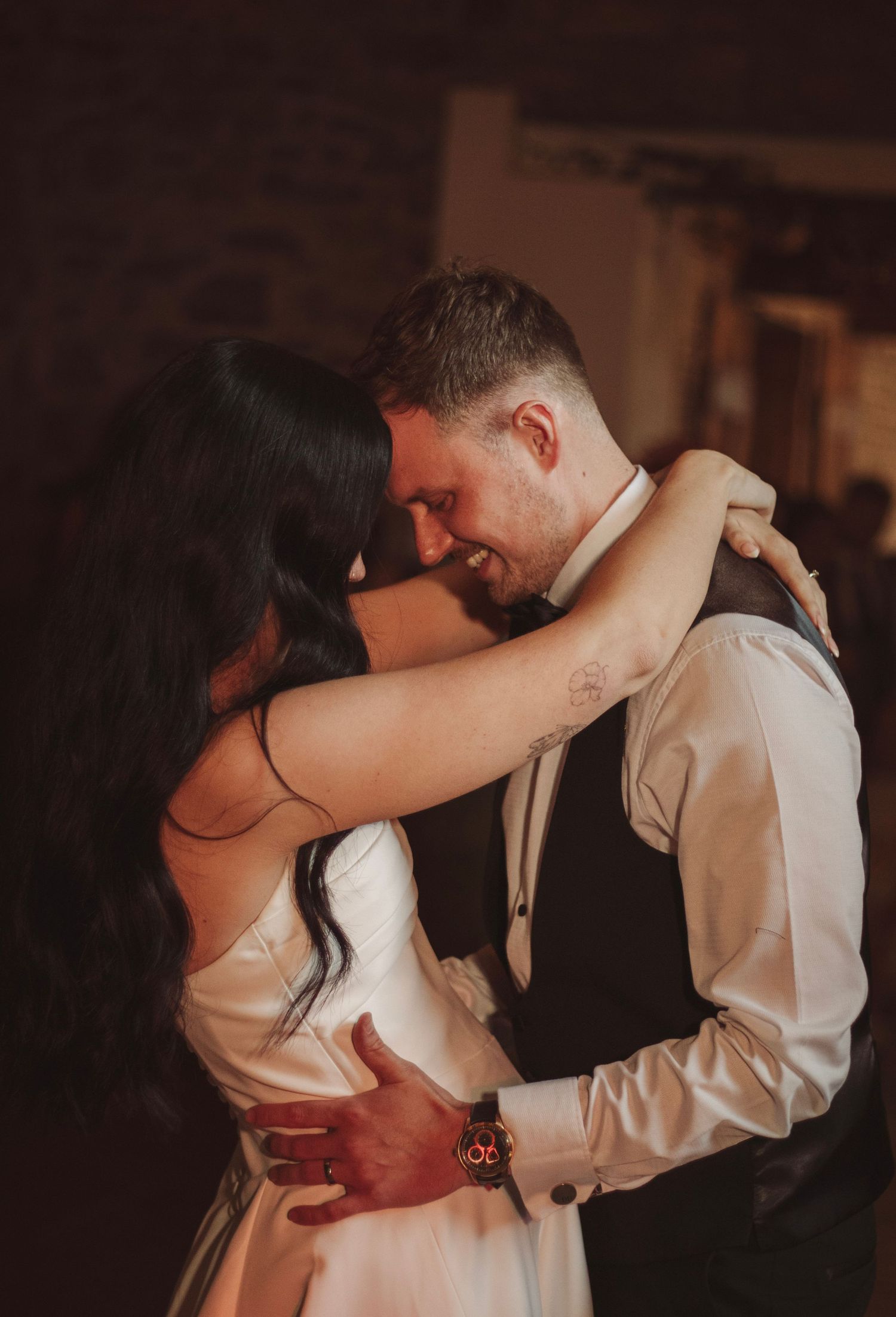 Bride in white dress and groom in vest share a tender first dance, embracing closely as he smiles in a warmly lit wedding venue.