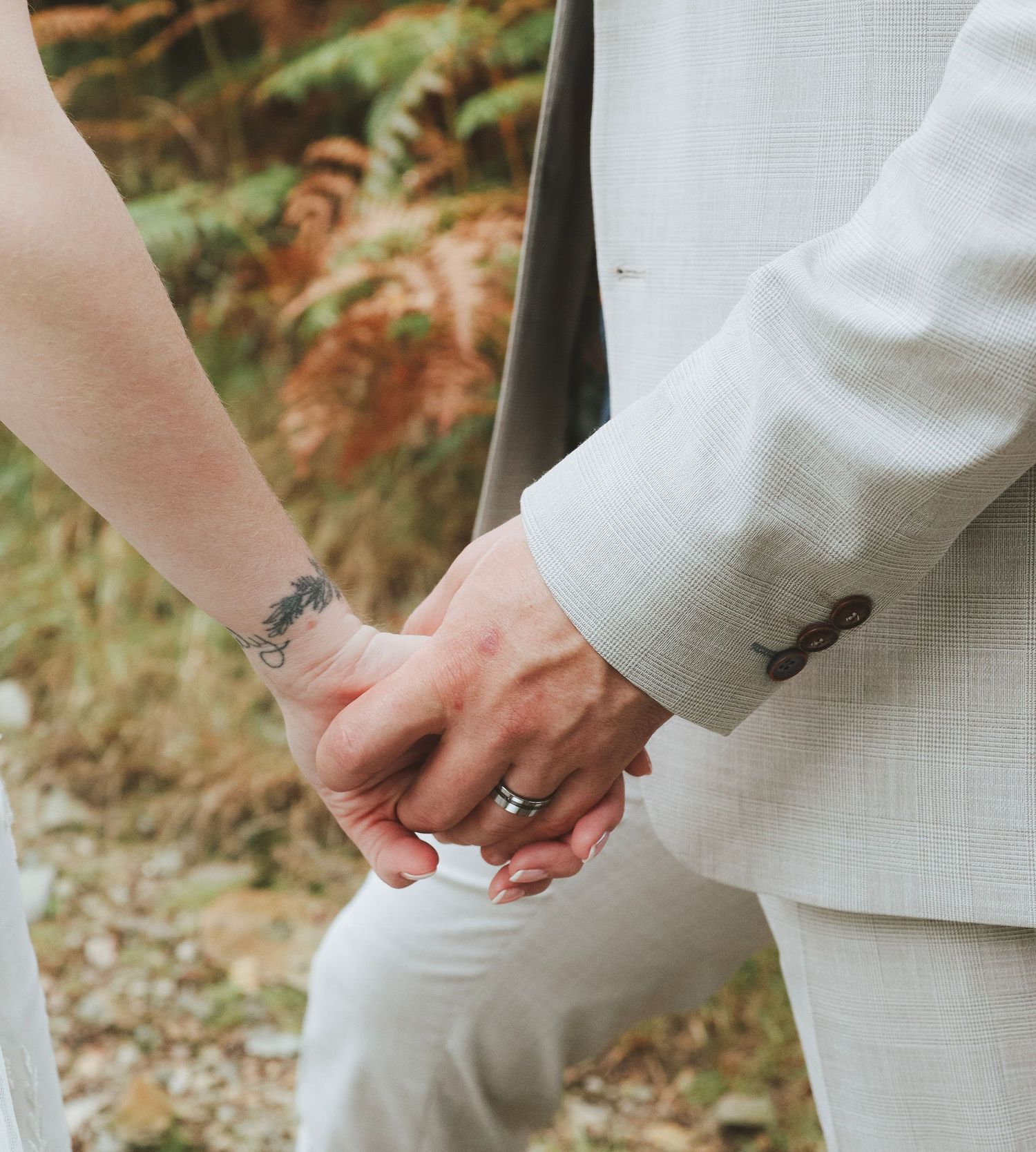 Couple holding hands outdoors, one wearing a light linen suit and a wedding ring, the other with a wrist tattoo, beside autumn ferns.