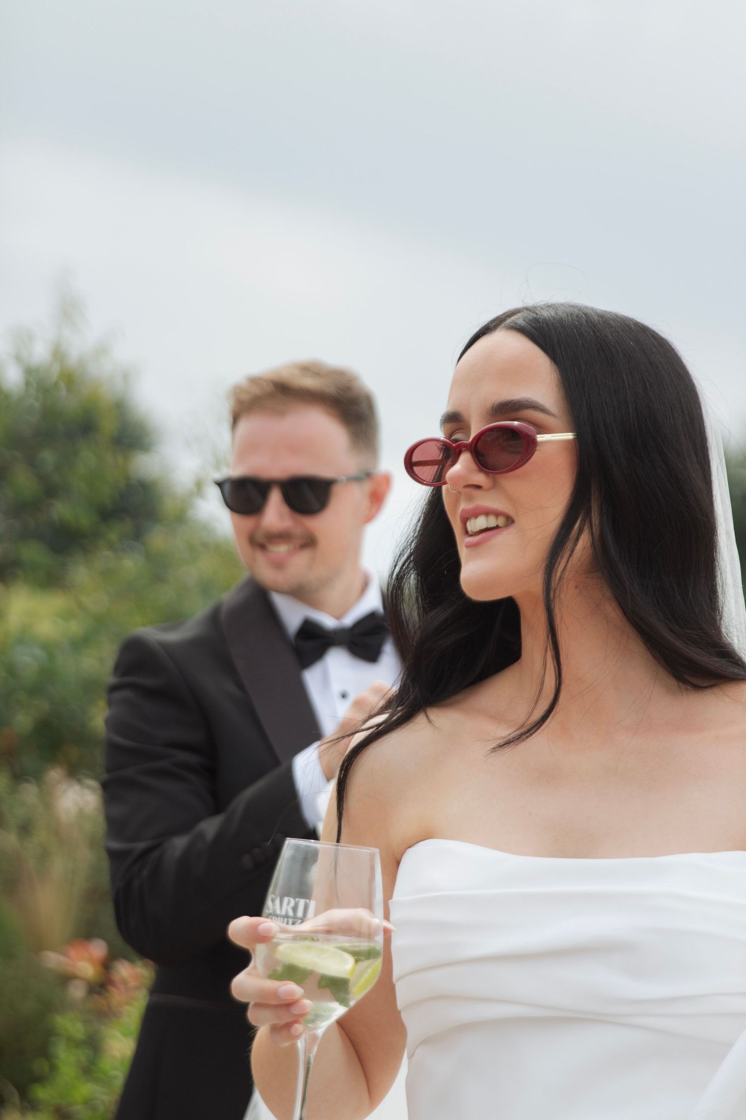 Bride in a white strapless dress and red sunglasses holds a glass of sparkling wine while a groom in a tuxedo smiles behind her outdoors.
