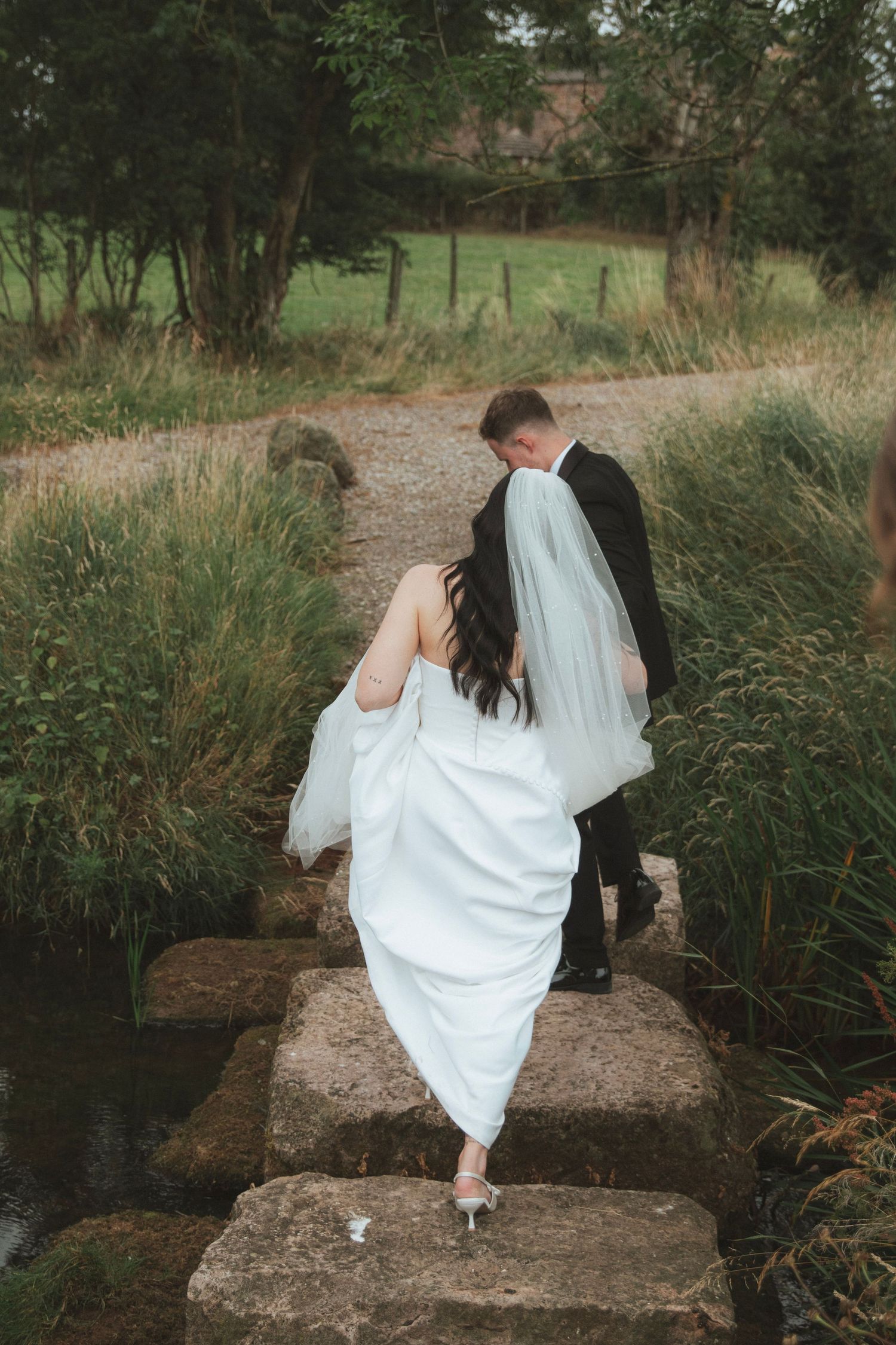 Bride in a white gown and veil crosses stone steps over a stream with her groom in a dark suit on a countryside path.