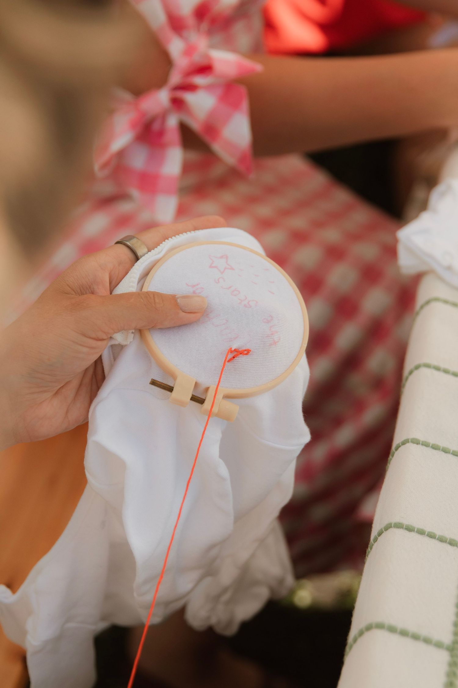 Close-up of hands doing embroidery with red thread on white fabric in a wooden hoop at an outdoor event.