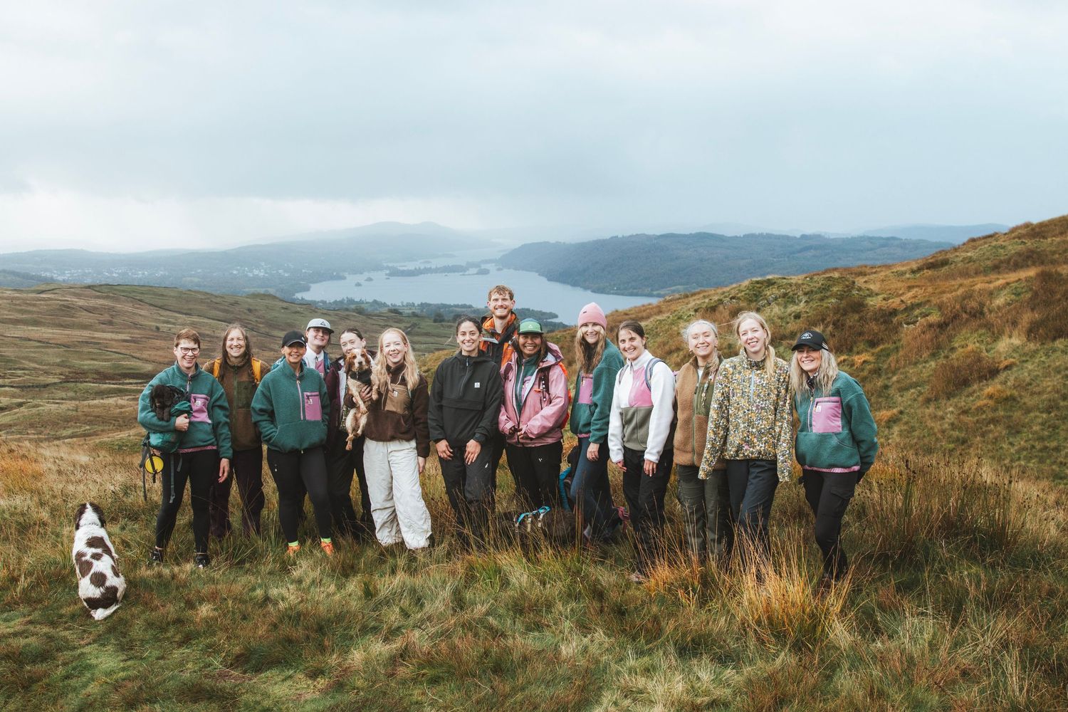 Group of hikers in jackets posing together on a grassy hillside with a misty lake and rolling mountains in the background.