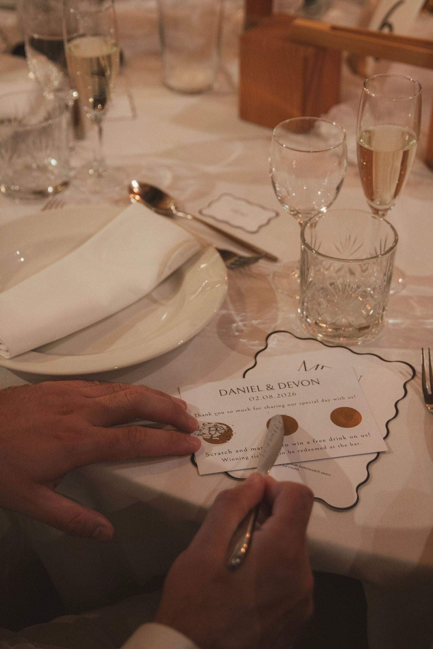 Guest scratches off a wedding game card for Daniel & Devon beside champagne flutes, glassware, and a folded napkin on a reception table.