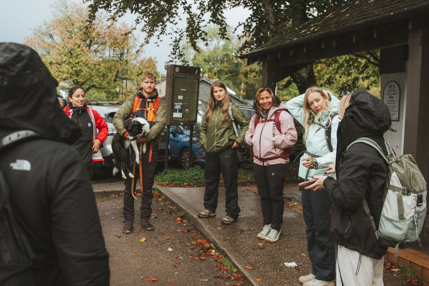 Group of hikers in jackets and backpacks gather outdoors near a wooden shelter as a guide wearing a camera speaks among autumn trees.