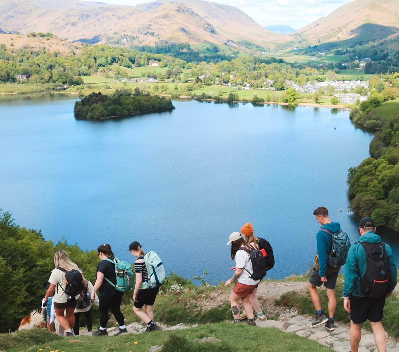 Group of hikers with backpacks descending a rocky trail overlooking a stunning blue lake surrounded by mountains.
