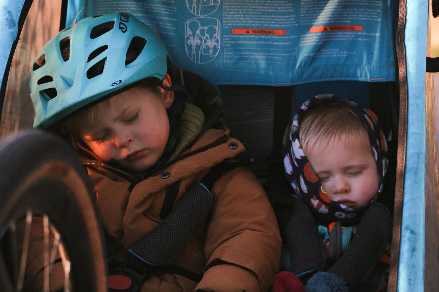 Two young children sleeping in a bike trailer, one wearing a teal helmet and the other in a red hat.