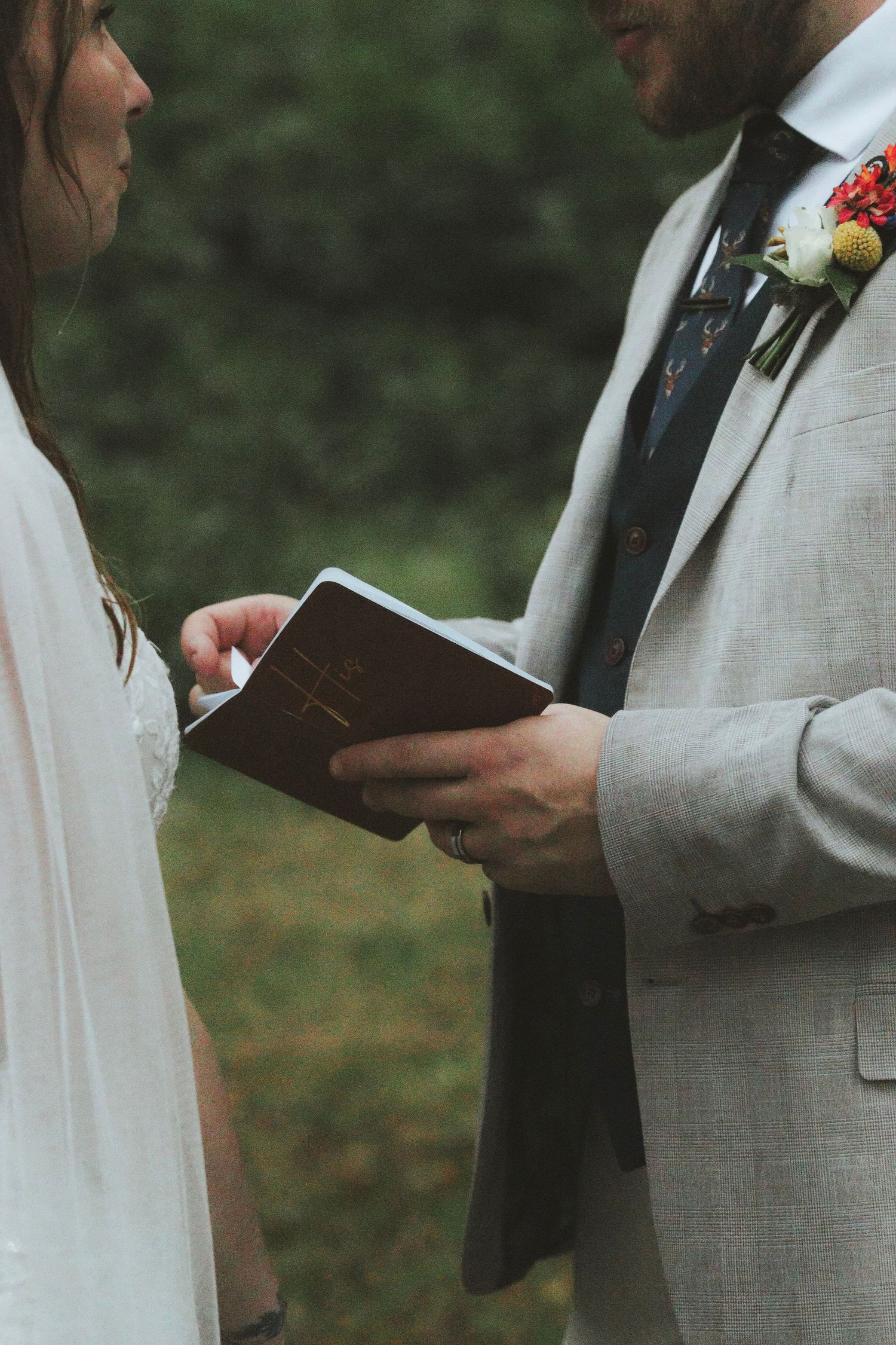 A groom in a gray suit with a floral boutonniere holds a small book while reading vows to his bride in an outdoor ceremony.