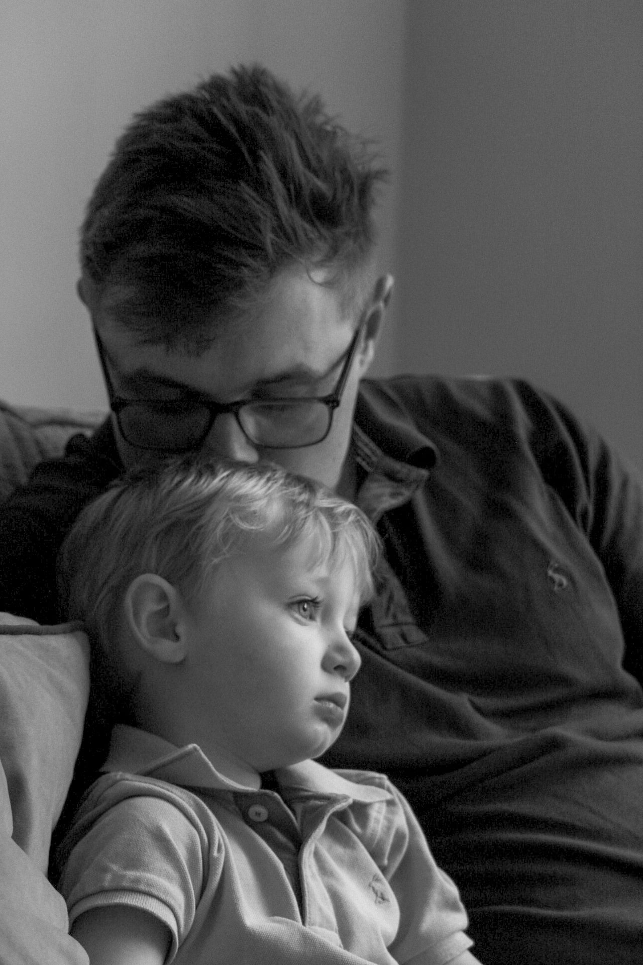 A father wearing glasses sits on a sofa with his young toddler son in a black and white portrait.