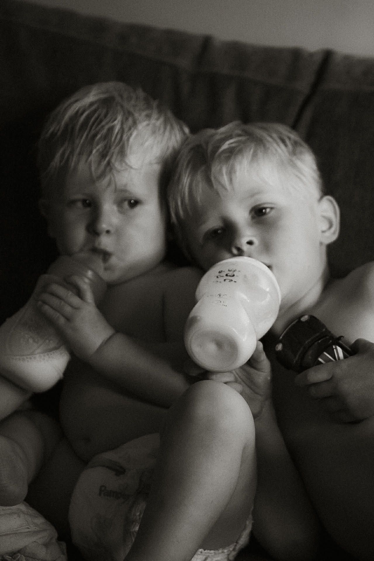 Black and white photo of two blonde toddlers drinking from baby bottles together.
