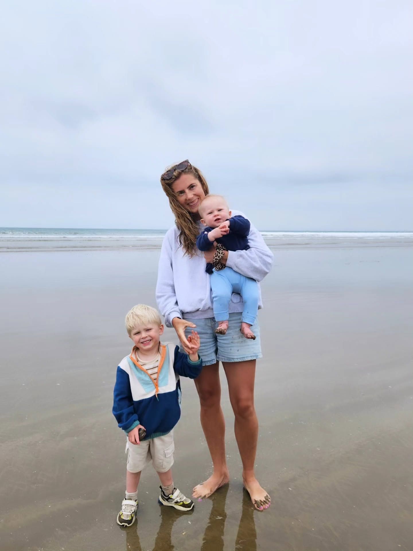 Woman holding baby with toddler beside her standing on a sandy beach on a cloudy day.