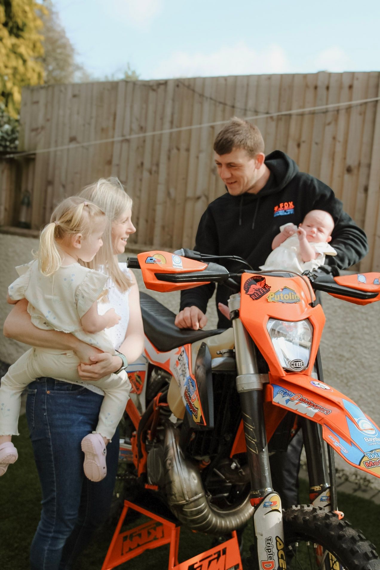 A young family with a toddler and newborn baby pose beside an orange KTM dirt bike outdoors.