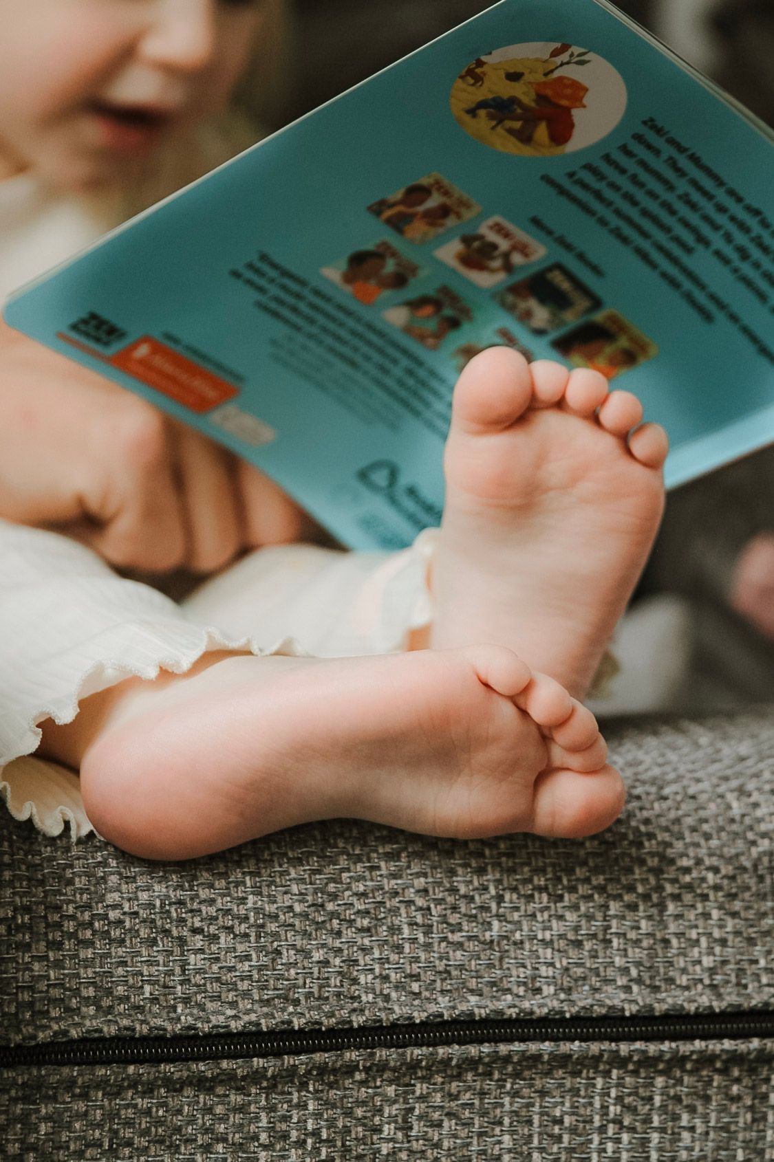 Close-up of a baby's tiny bare feet resting on a sofa while holding a colorful picture book.