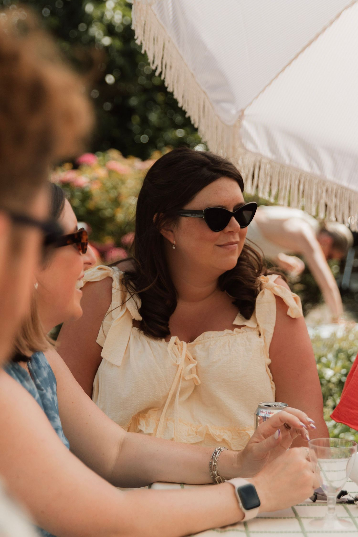 Woman in sunglasses and a cream ruffled top sits at an outdoor table under a fringed umbrella, chatting with friends in a sunny garden.