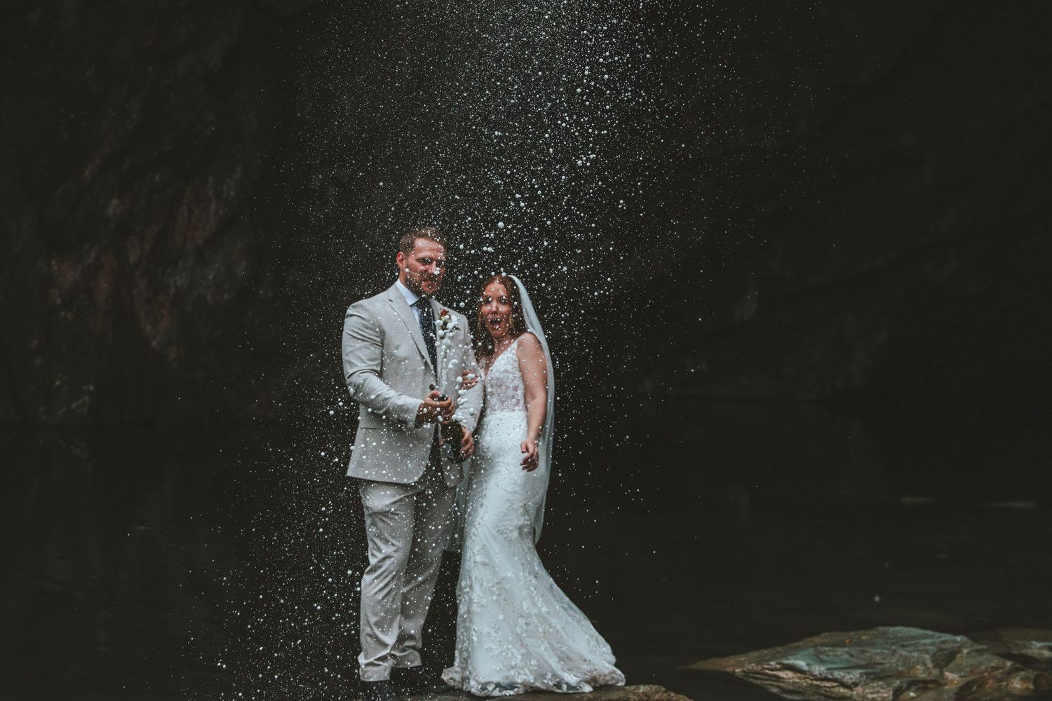 Bride in a white lace gown and groom in a light gray suit pose together beneath a misty waterfall spray in a dark, rocky outdoor setting.