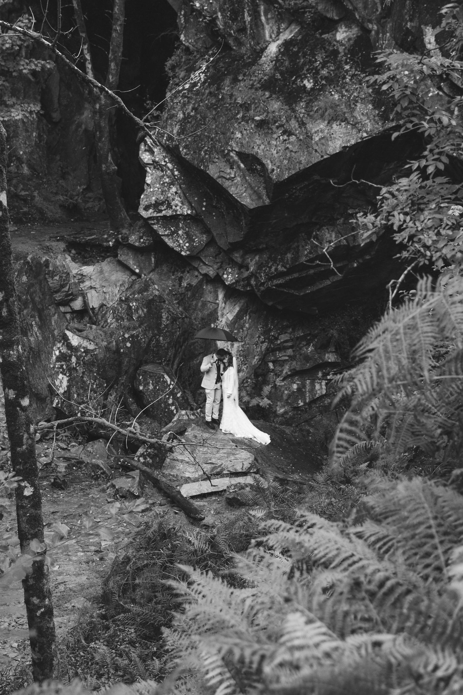 Black and white photo of a bride and groom sharing an umbrella beneath a rocky cliff overhang, surrounded by ferns in a forest.