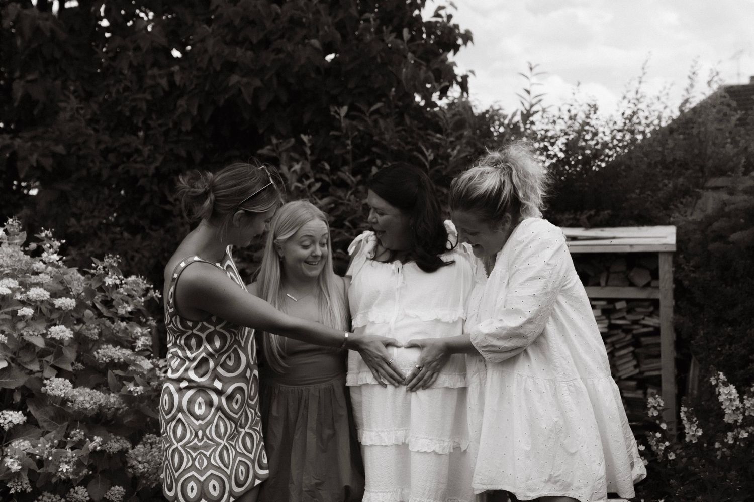 Black and white photo of four women gathered in a garden touching a pregnant belly joyfully.