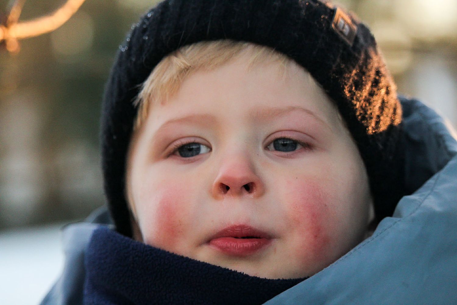 Close-up portrait of a young child with rosy cheeks wearing a dark knit hat outdoors in winter.