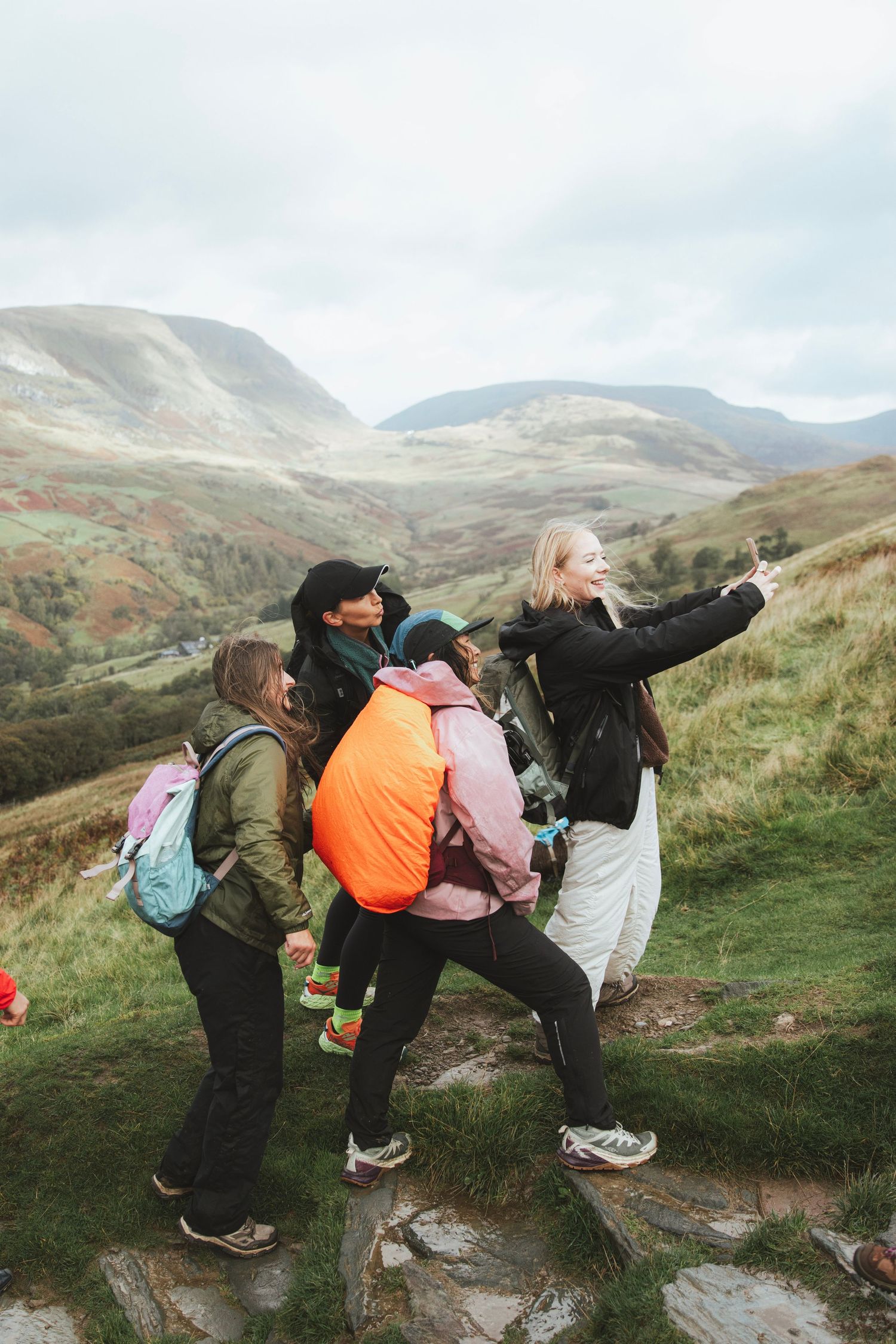 Group of hikers taking a selfie on a rocky mountain trail with green hills and valley views under a cloudy sky.