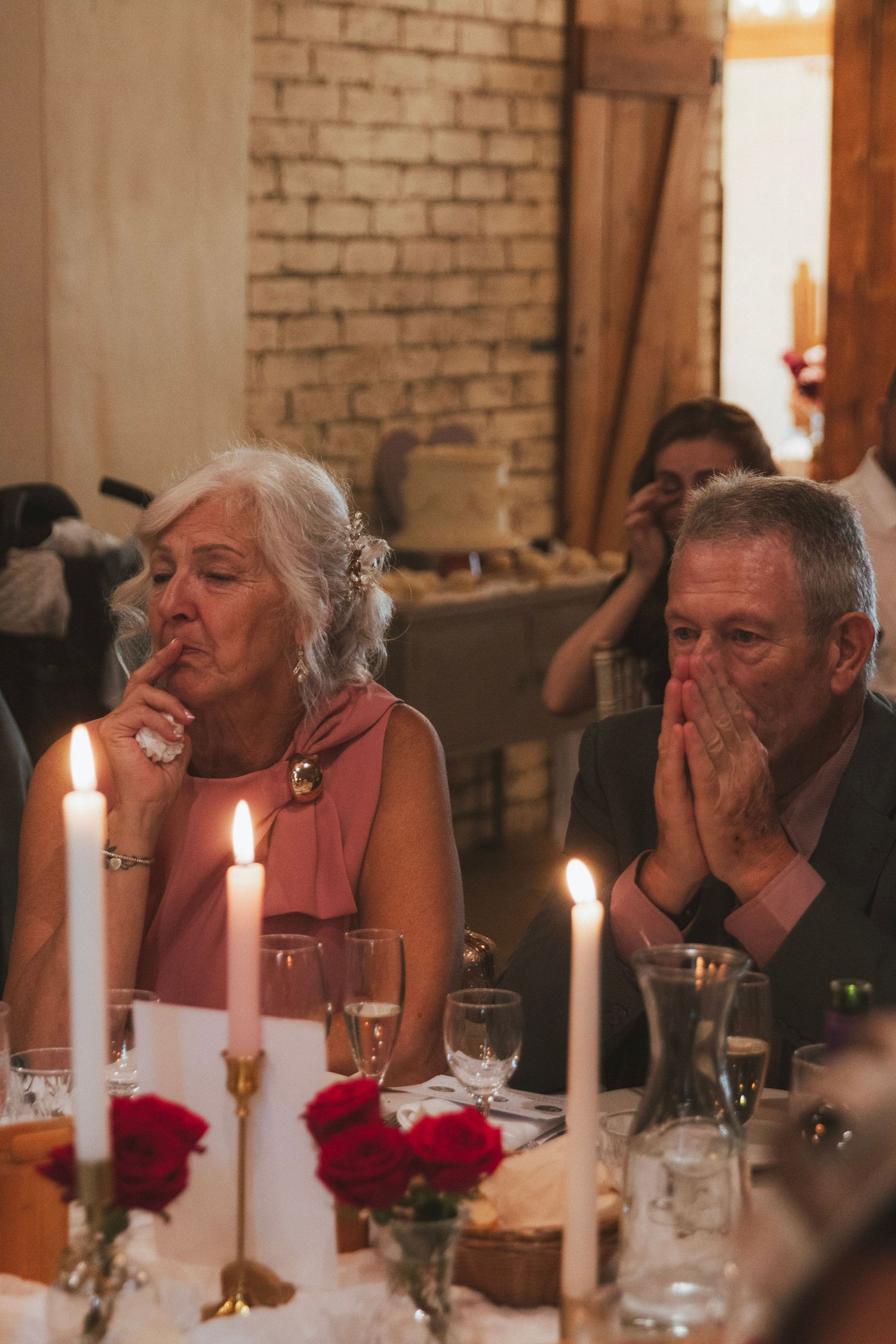 Two emotional wedding guests at a candlelit reception table, one dabbing her eyes and the other covering his mouth with his hands.