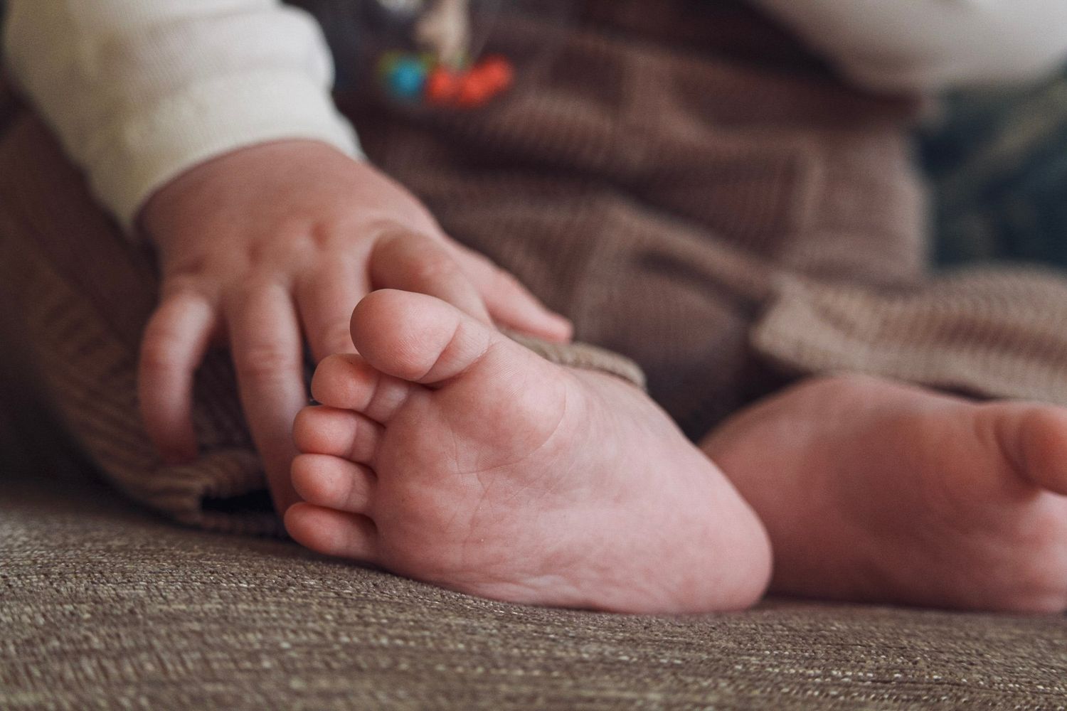 A close-up of a baby's tiny foot and hand resting gently on a soft brown surface.