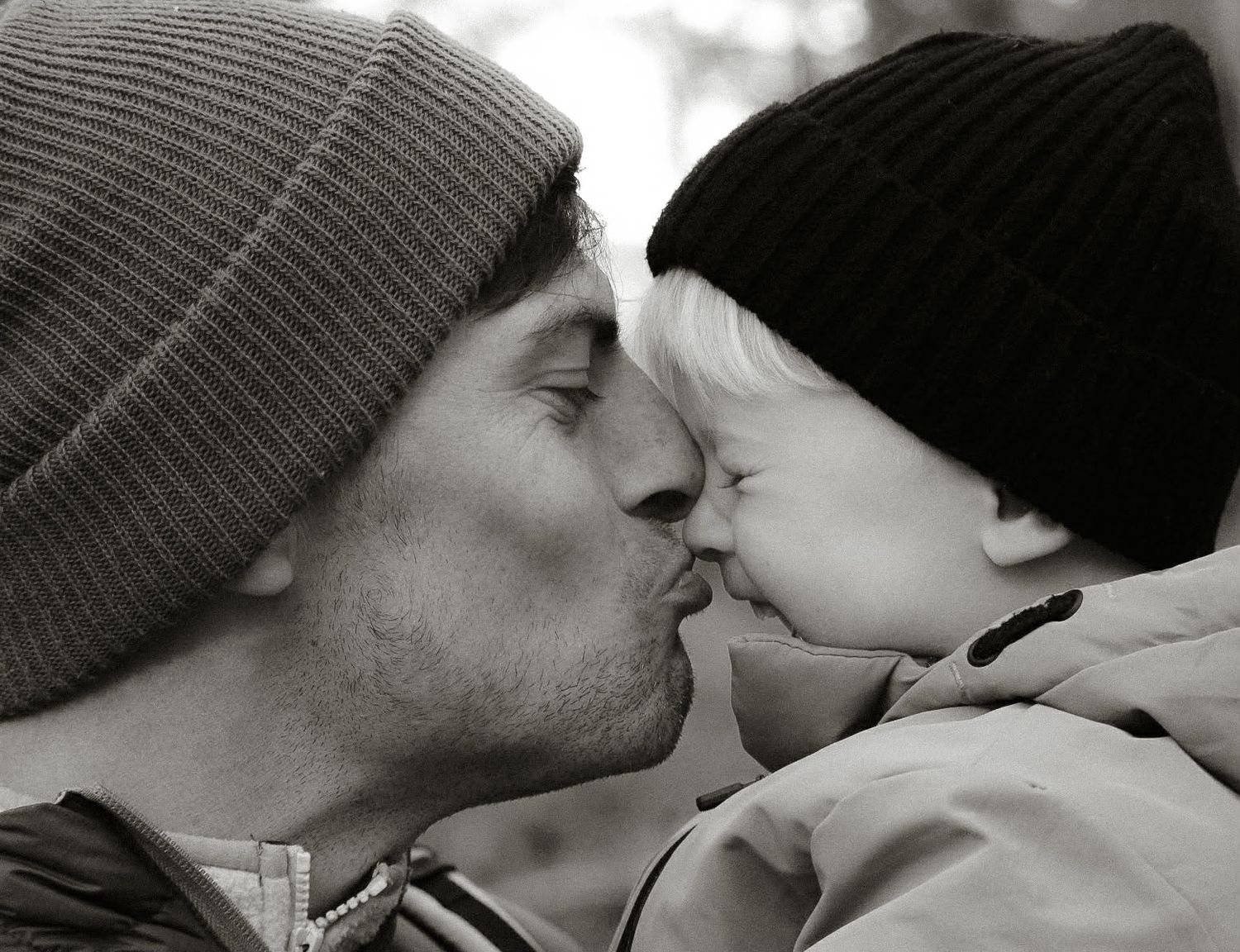 Black and white photo of a father in a beanie hat gently kissing his baby outdoors.