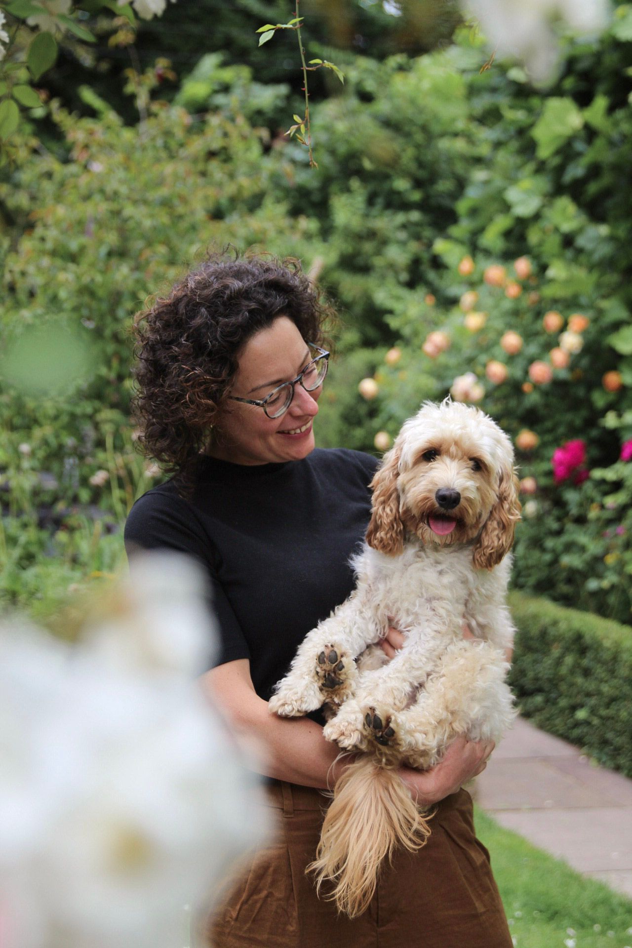 Smiling woman with curly hair holding a fluffy Cockapoo dog in a garden.