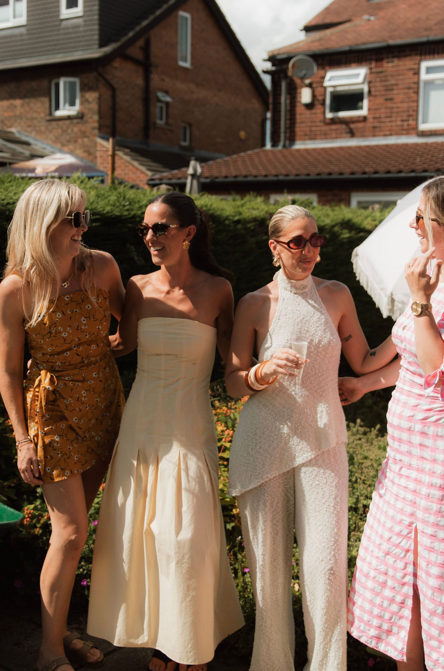 Four stylish women in summer dresses laugh and chat together at an outdoor garden party.