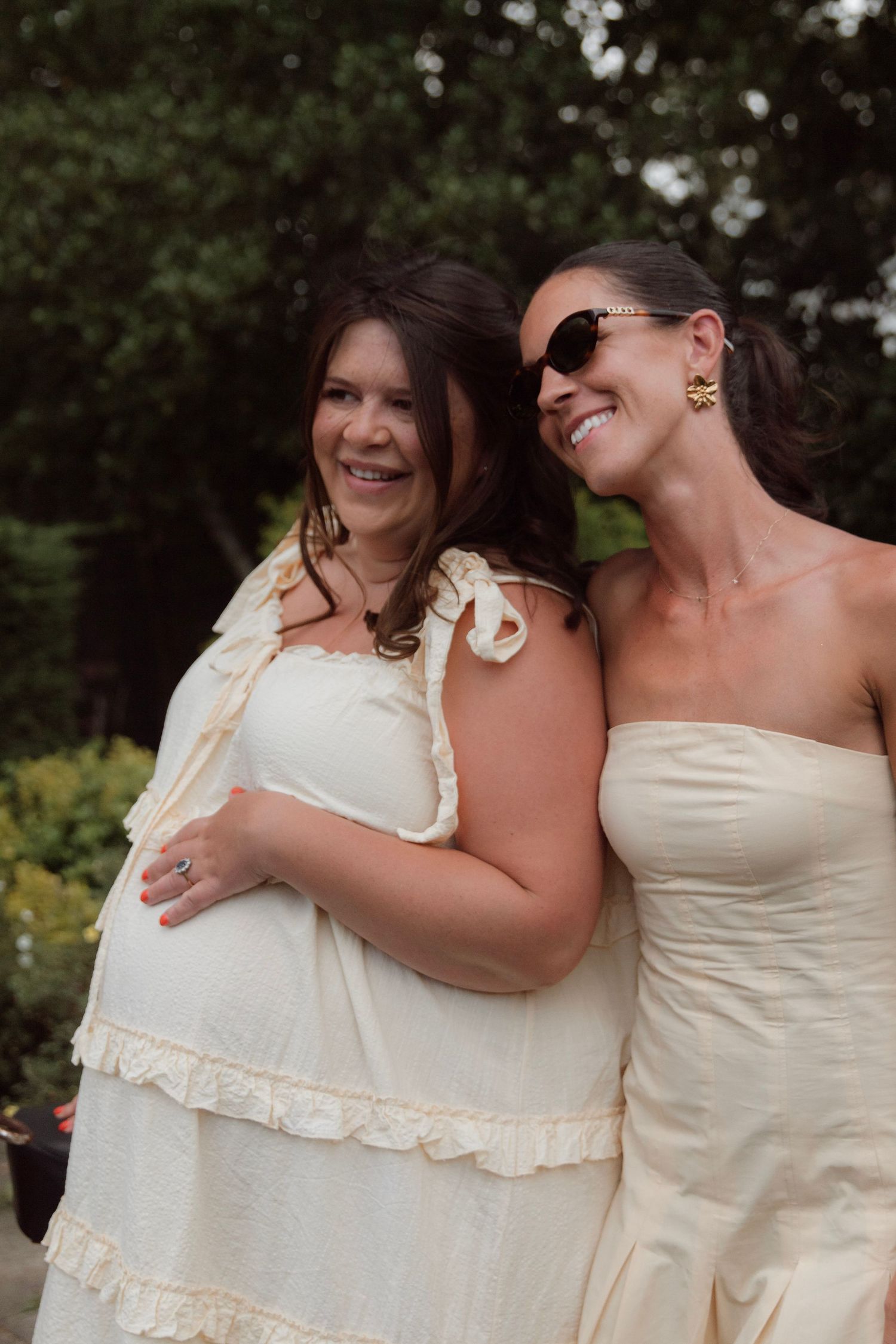 Two smiling women in cream dresses pose together outdoors in a garden setting on a sunny day.