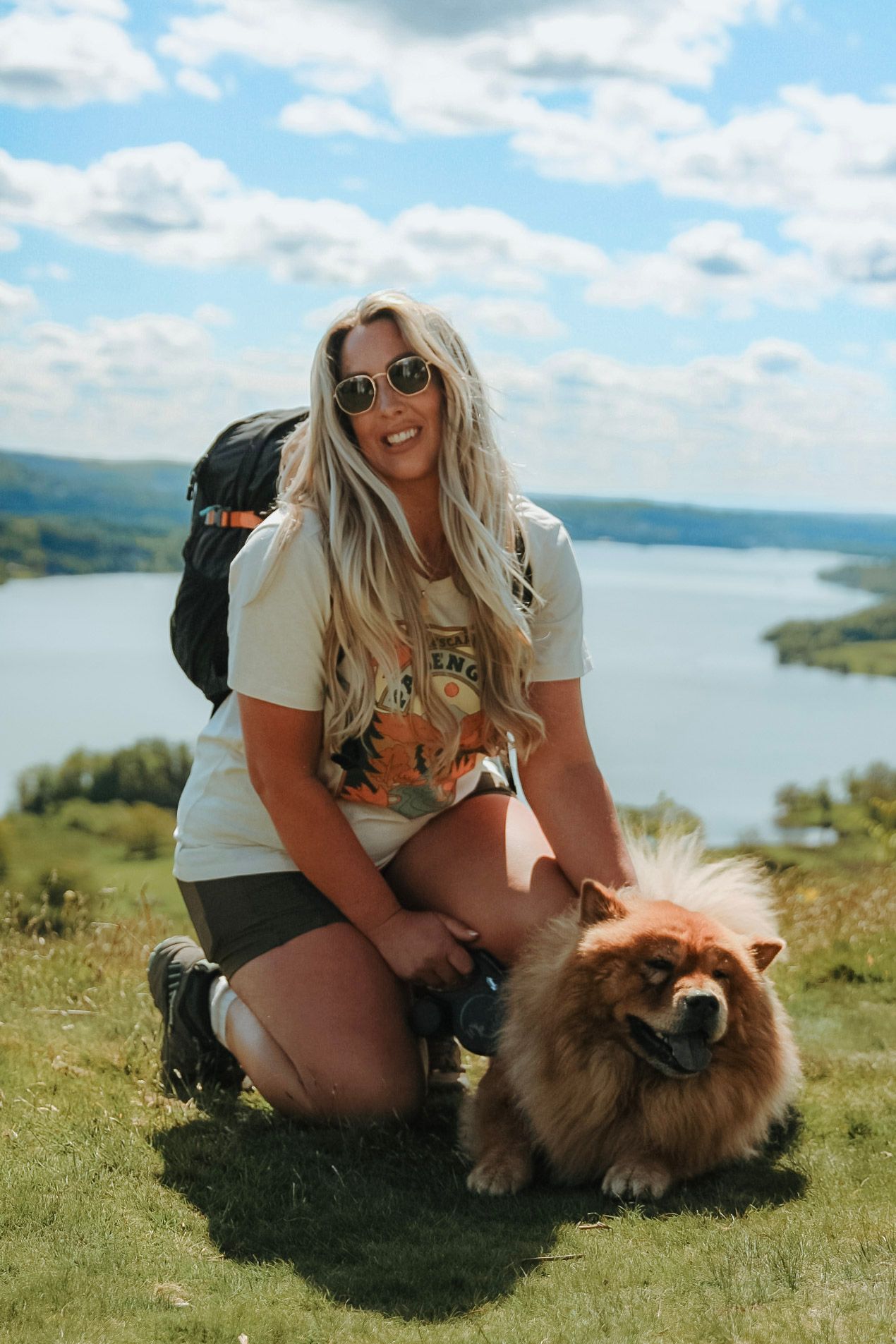 Smiling blonde woman with backpack kneeling beside a fluffy Chow Chow dog on a hilltop with lake views.