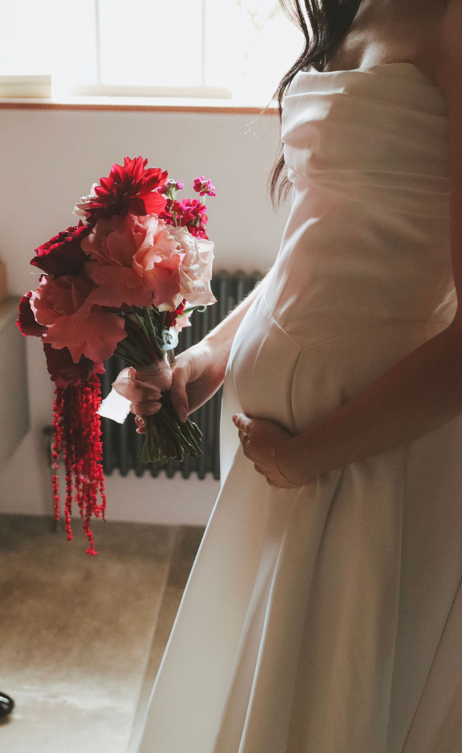 Bride in a strapless white satin gown holds a vibrant bouquet of pink and red peonies, dahlias and trailing amaranthus beside a window.