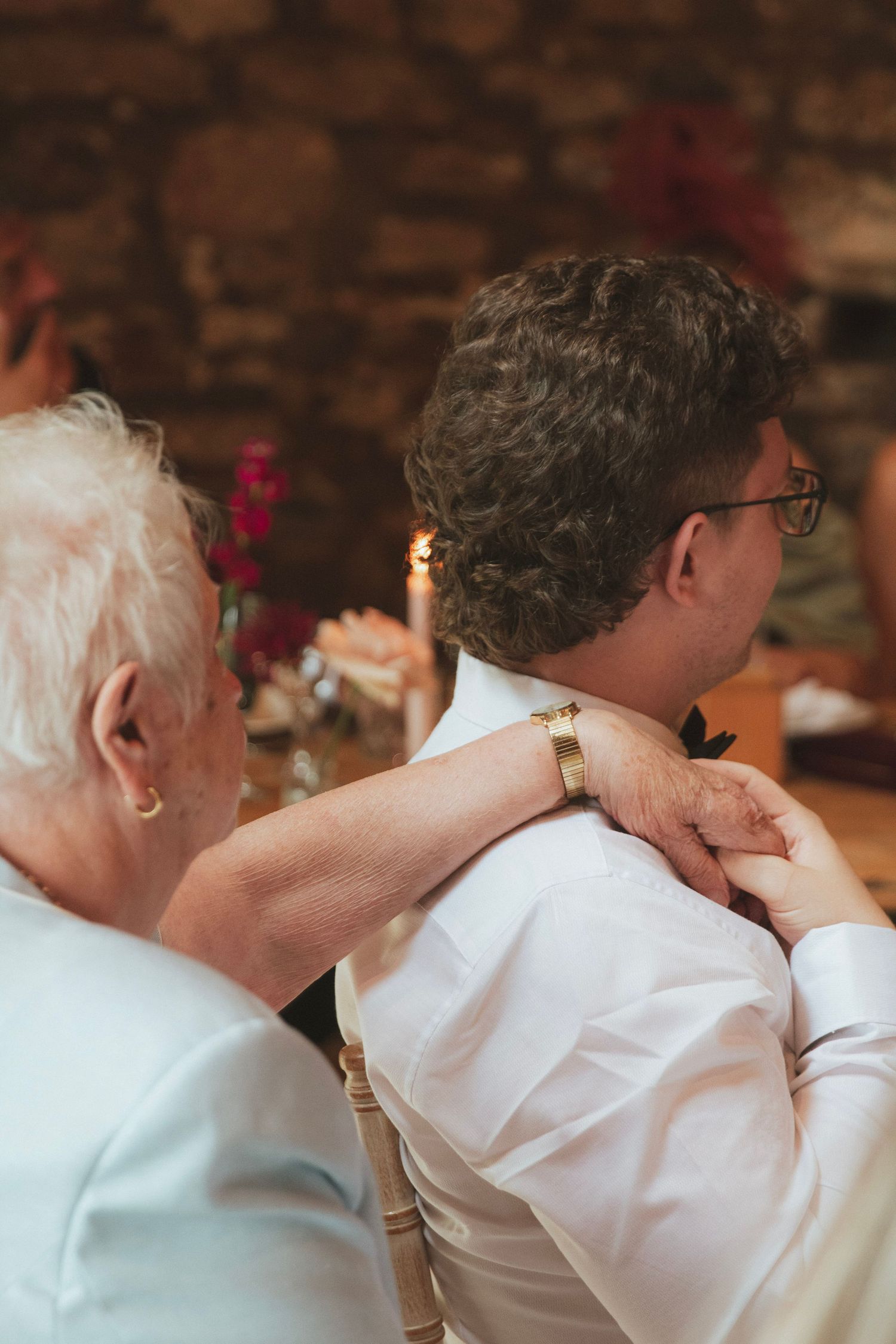 Elderly woman rests her hand affectionately on the shoulder of a seated man in glasses and a white shirt at a candlelit reception table.