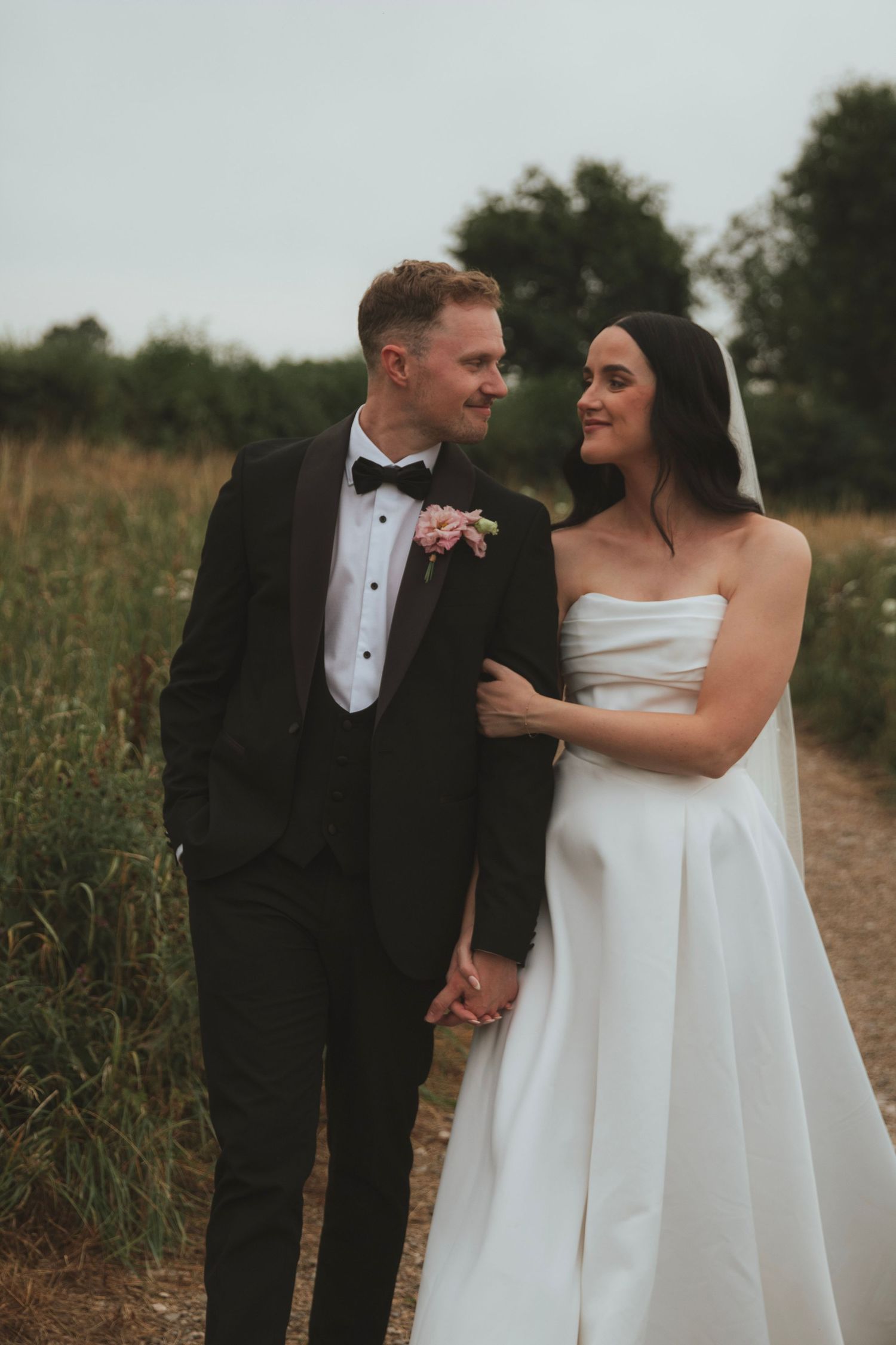 A bride and groom hold hands on a country path, smiling at each other, dressed in a white gown and black tuxedo.