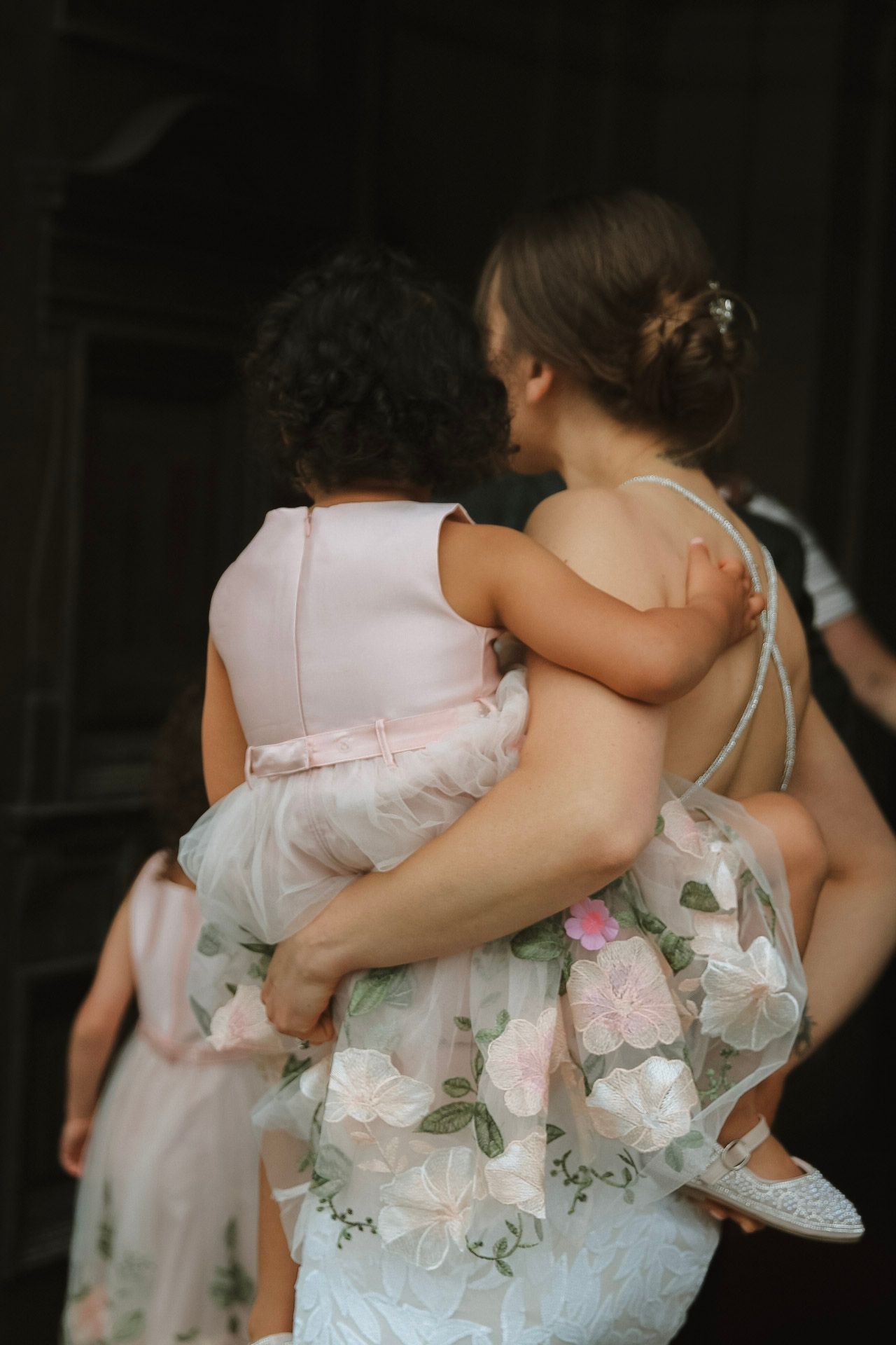 A woman in a floral wedding dress holds a young girl in a pink dress at a wedding celebration.