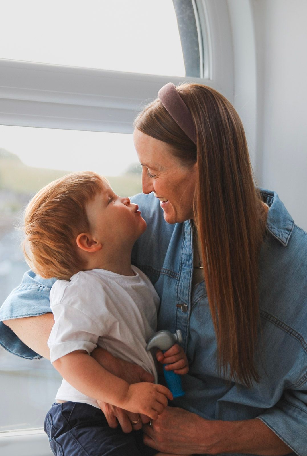 A mother with long hair and denim jacket smiling at her laughing toddler son by a window.